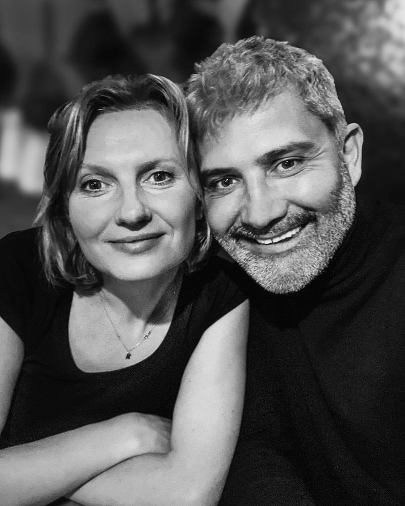 A black and white portrait of a couple smiling together, with the person on the right wearing a dark sweater.