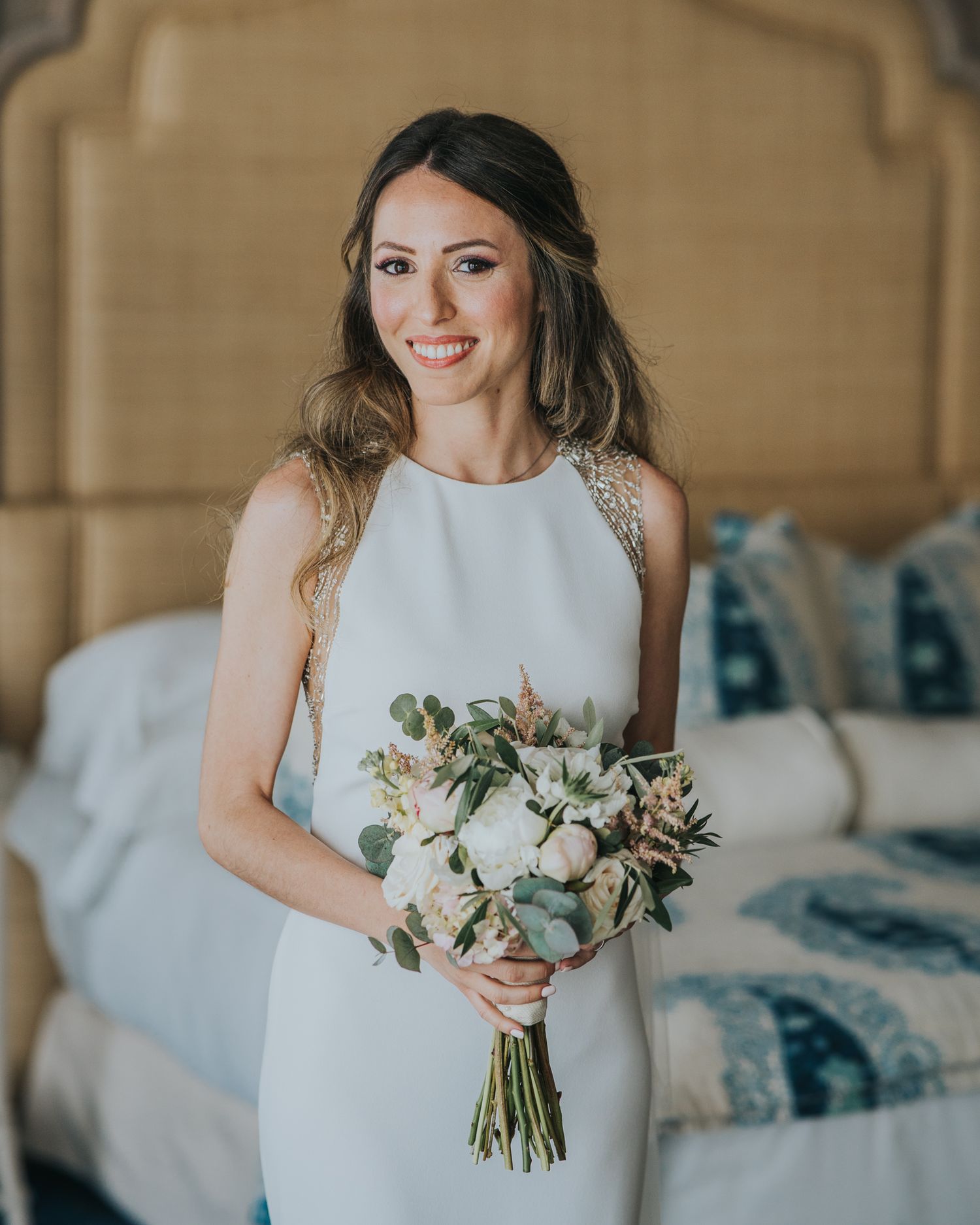 A person in a sleeveless white dress holds an elegant bouquet of white and green flowers in a bedroom setting.