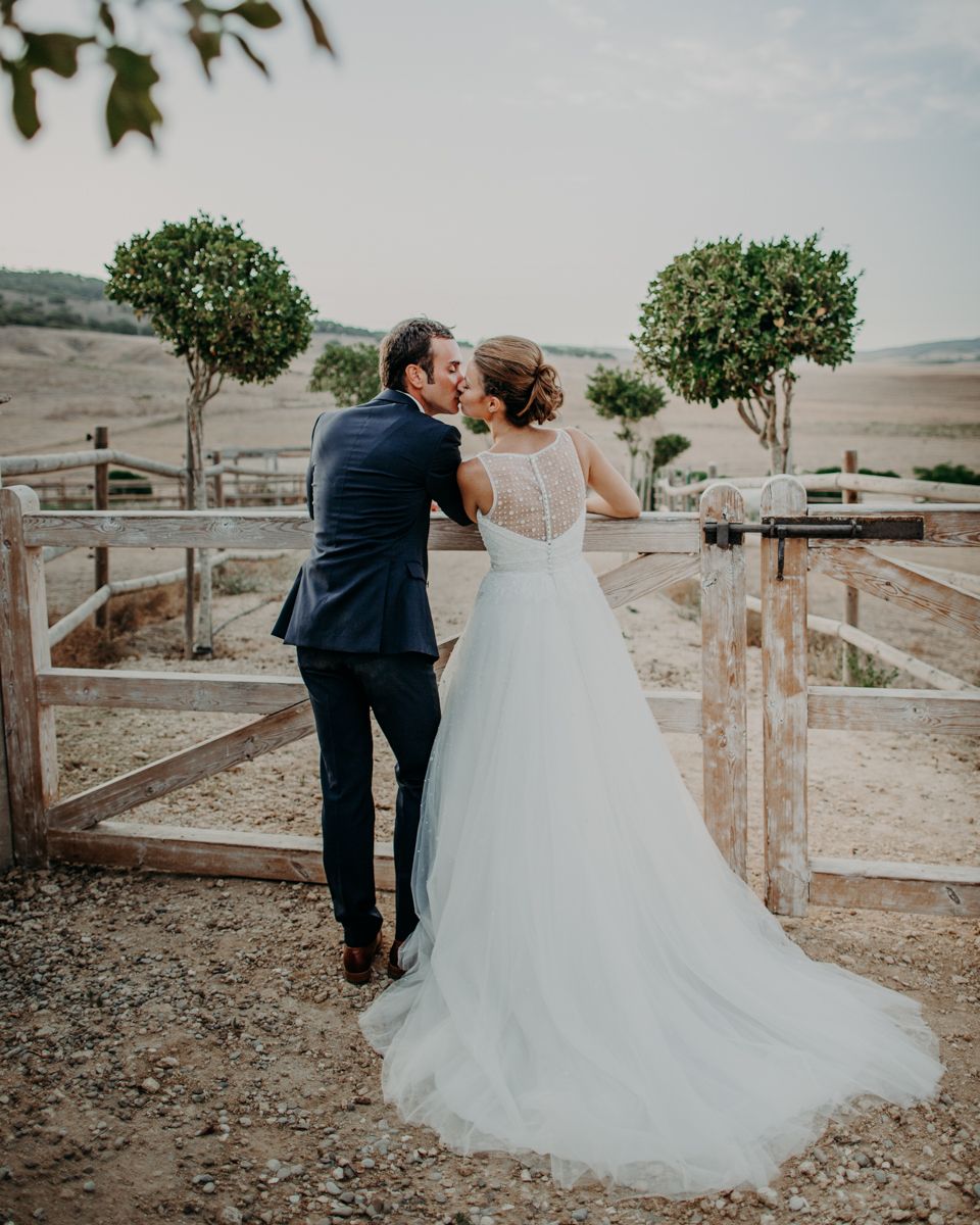 A romantic newlywed couple sharing an intimate moment by a rustic fence overlooking rolling countryside terrain.