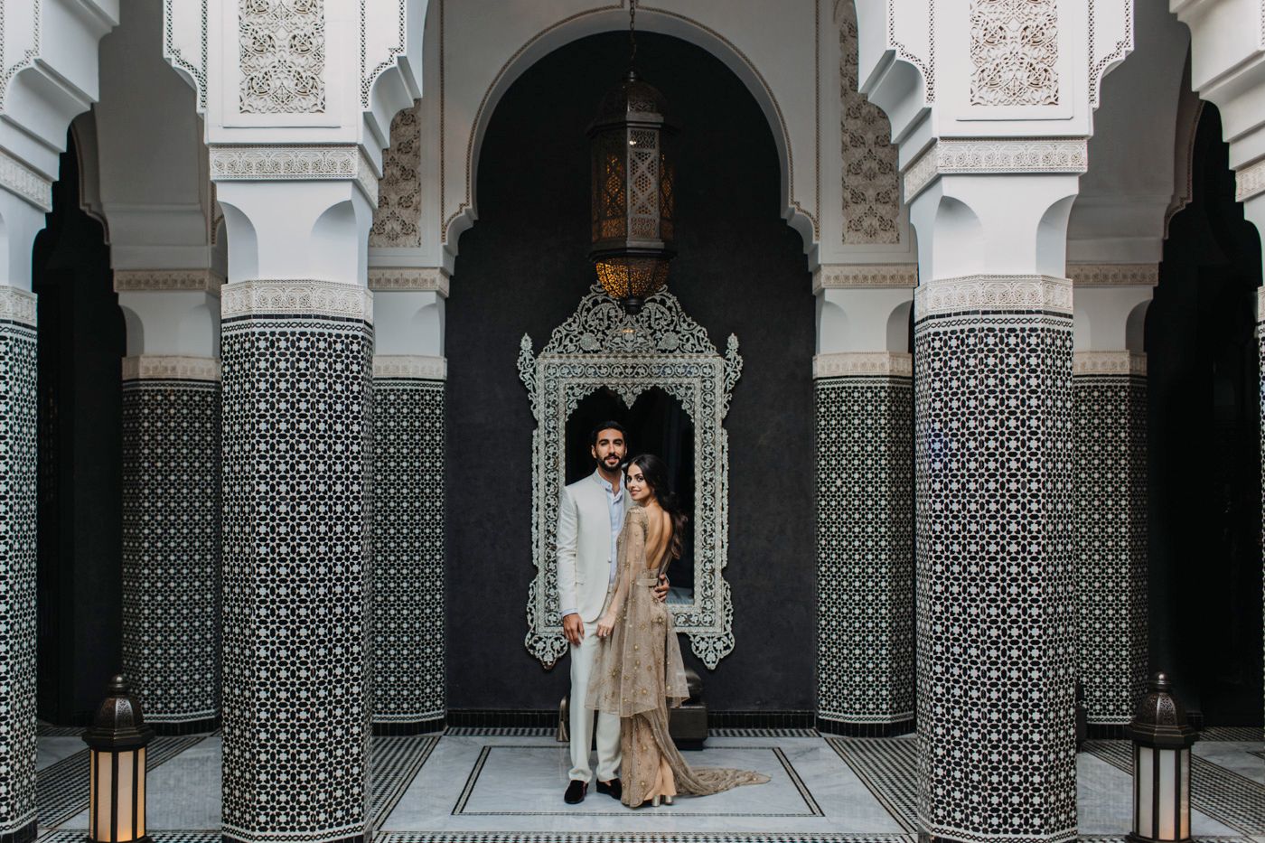 Elegant couple stands in doorway of Moroccan-inspired architecture with detailed tile patterns.