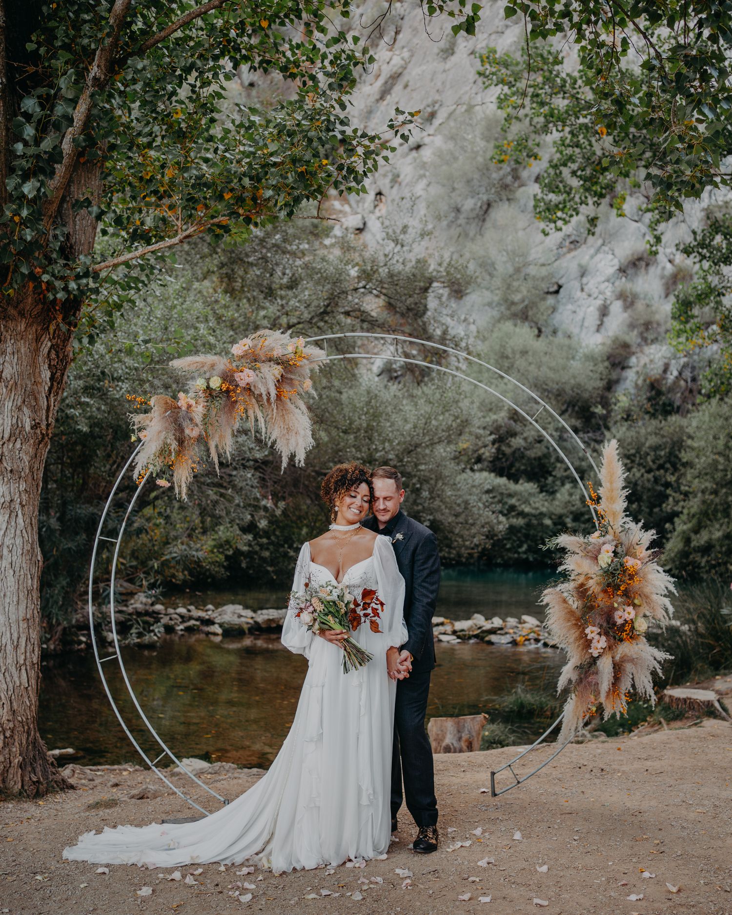Wedding couple poses under circular arch with pampas grass decor by river and trees.