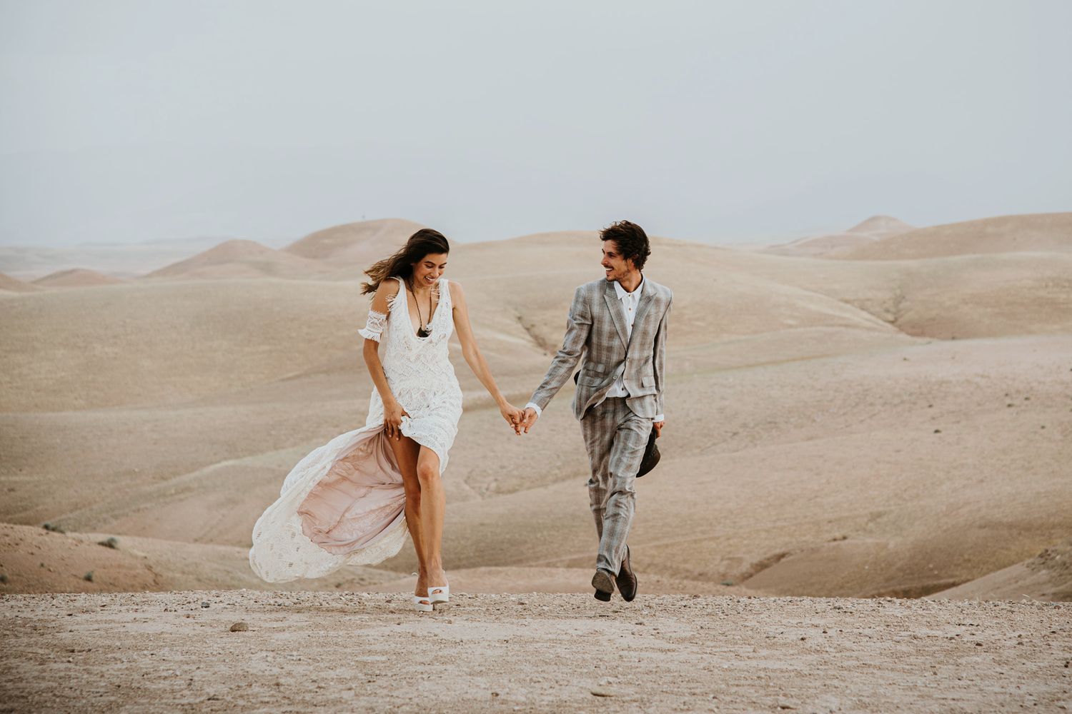 Couple holds hands while walking through desert landscape in wedding attire.
