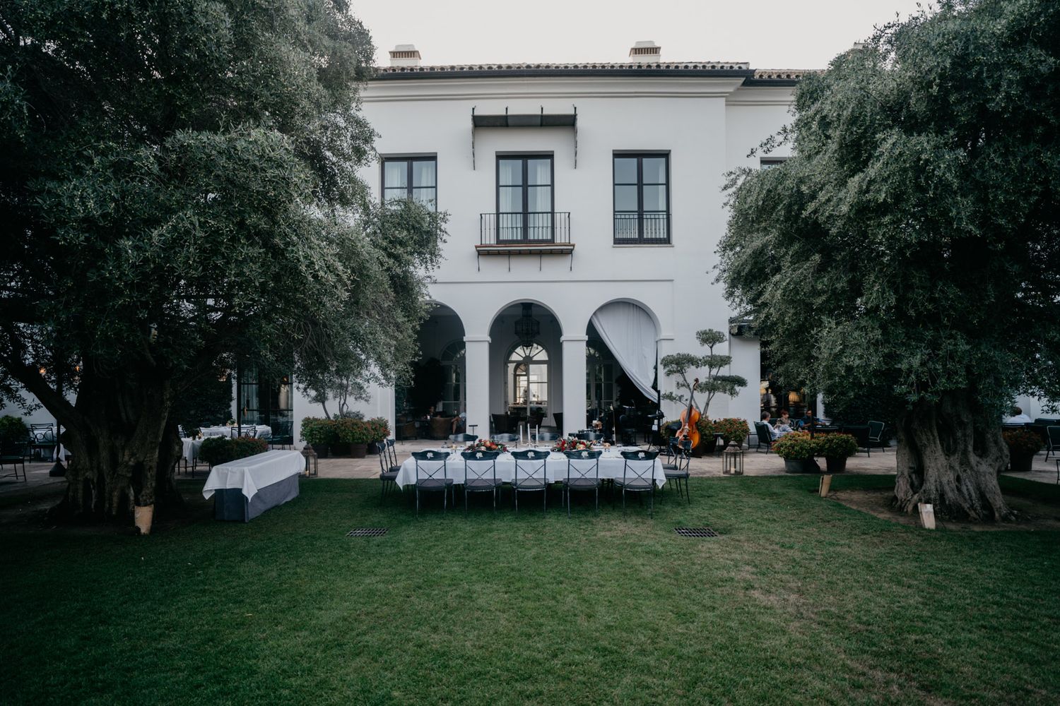 Elegant outdoor wedding reception setup with white tables in front of a Mediterranean-style villa between two mature trees.