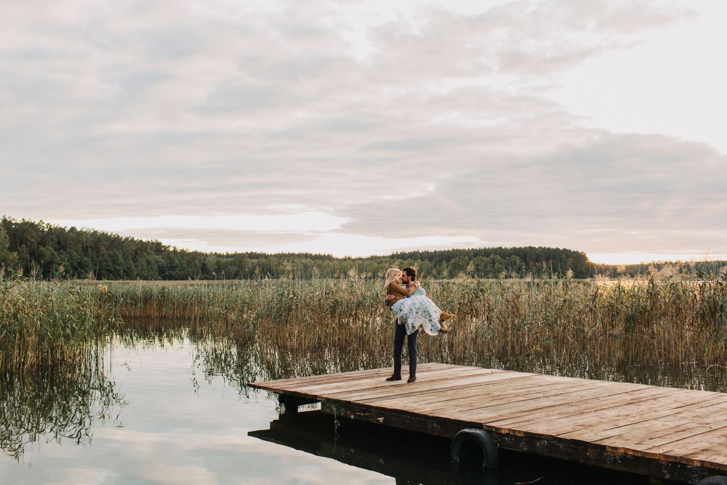 A silhouette stands on a wooden dock overlooking a tranquil lake surrounded by tall reeds at sunset.