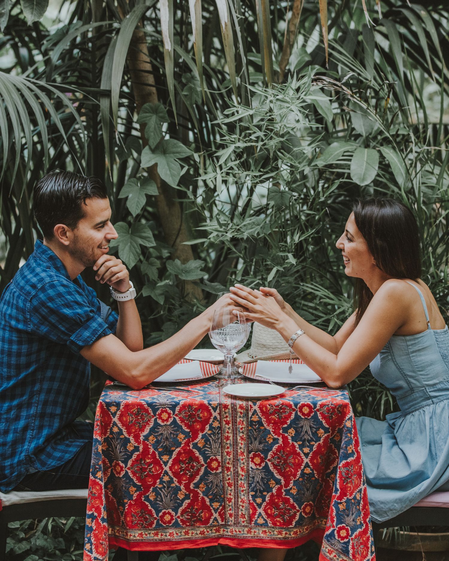 A couple enjoying an intimate dinner date at a table with a decorative red patterned tablecloth surrounded by tropical plants.