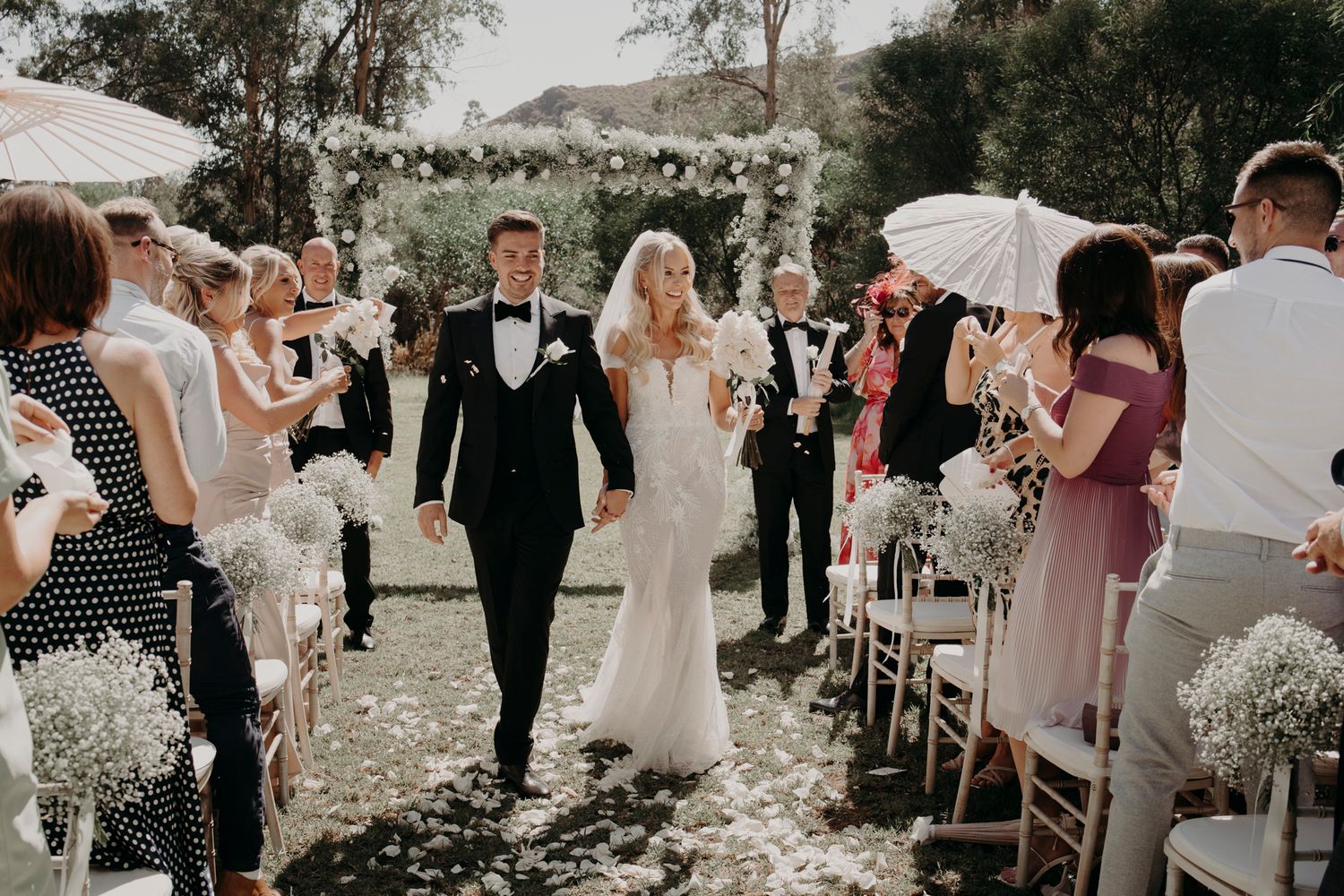 A joyful outdoor wedding ceremony with guests lining the aisle as couple walks between white floral arrangements and guests.