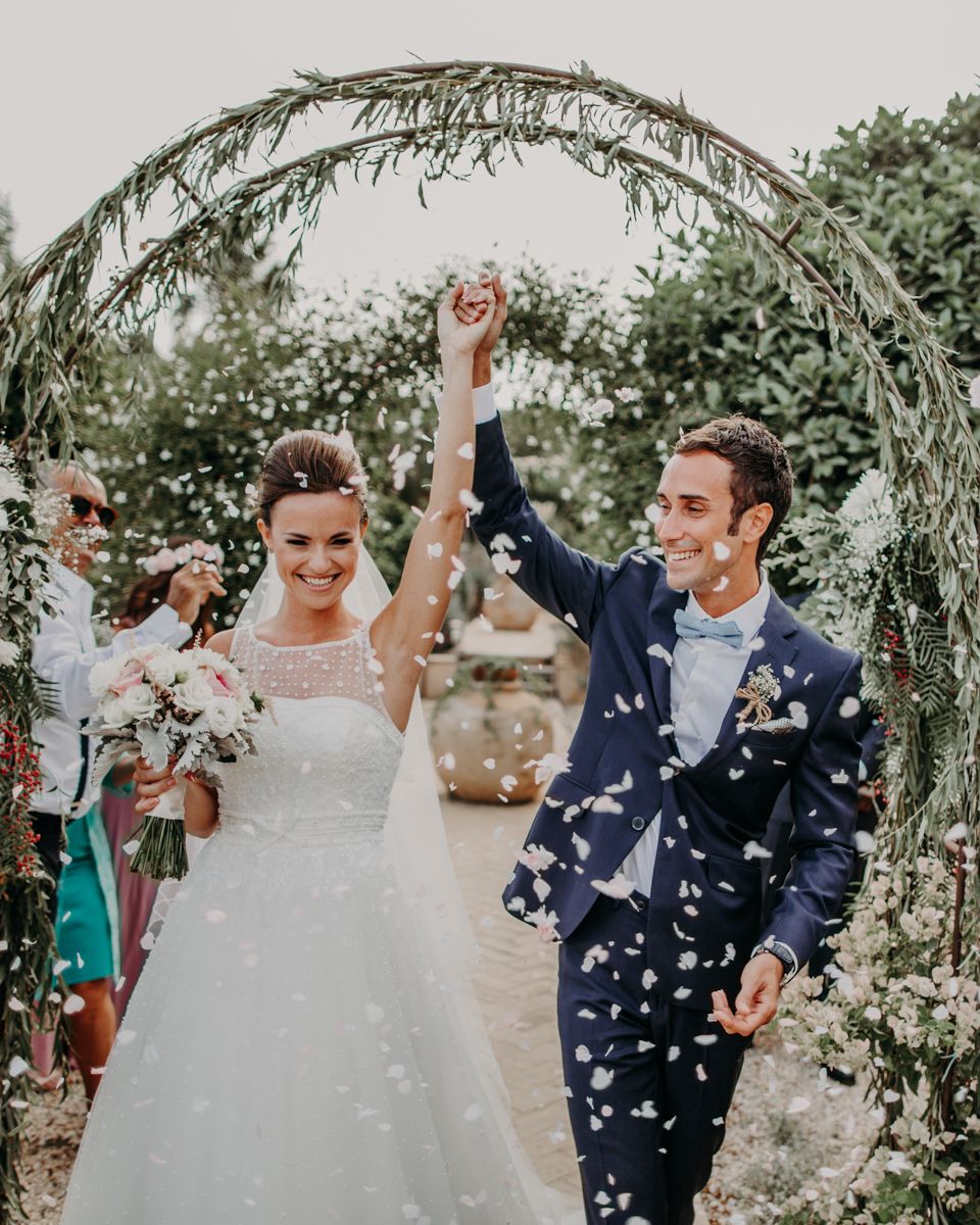 Newlyweds celebrate under a rustic greenery arch as confetti flutters around them during an outdoor garden ceremony.