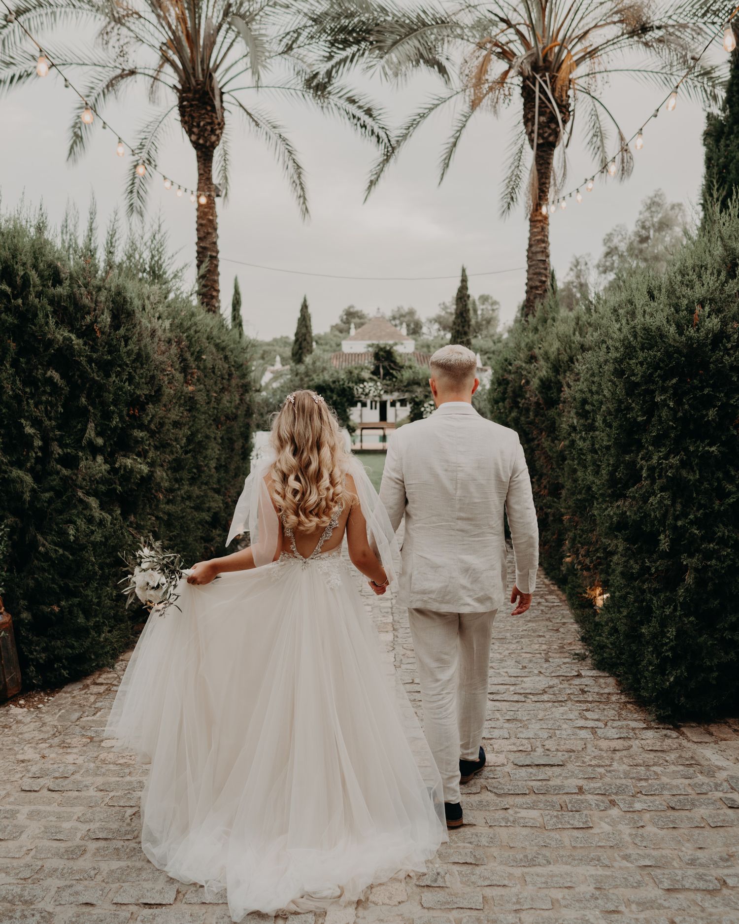 A wedding couple walks down a palm-lined garden path in elegant attire.