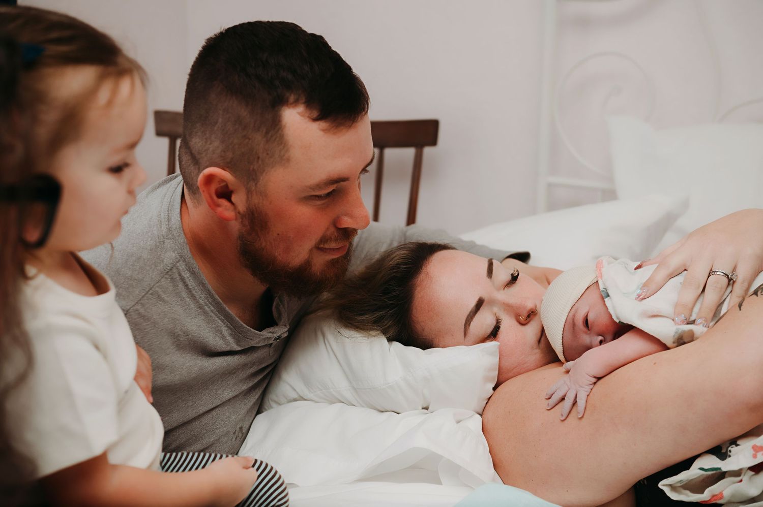 A family gathers on a bed as a mother holds her newborn baby while a toddler and father look on lovingly.