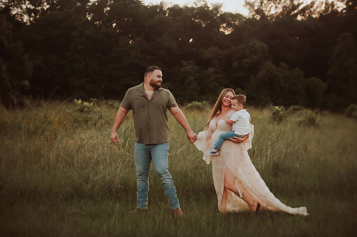 Family of three posing in a lush green meadow at golden hour, parents holding hands while mom cradles toddler.