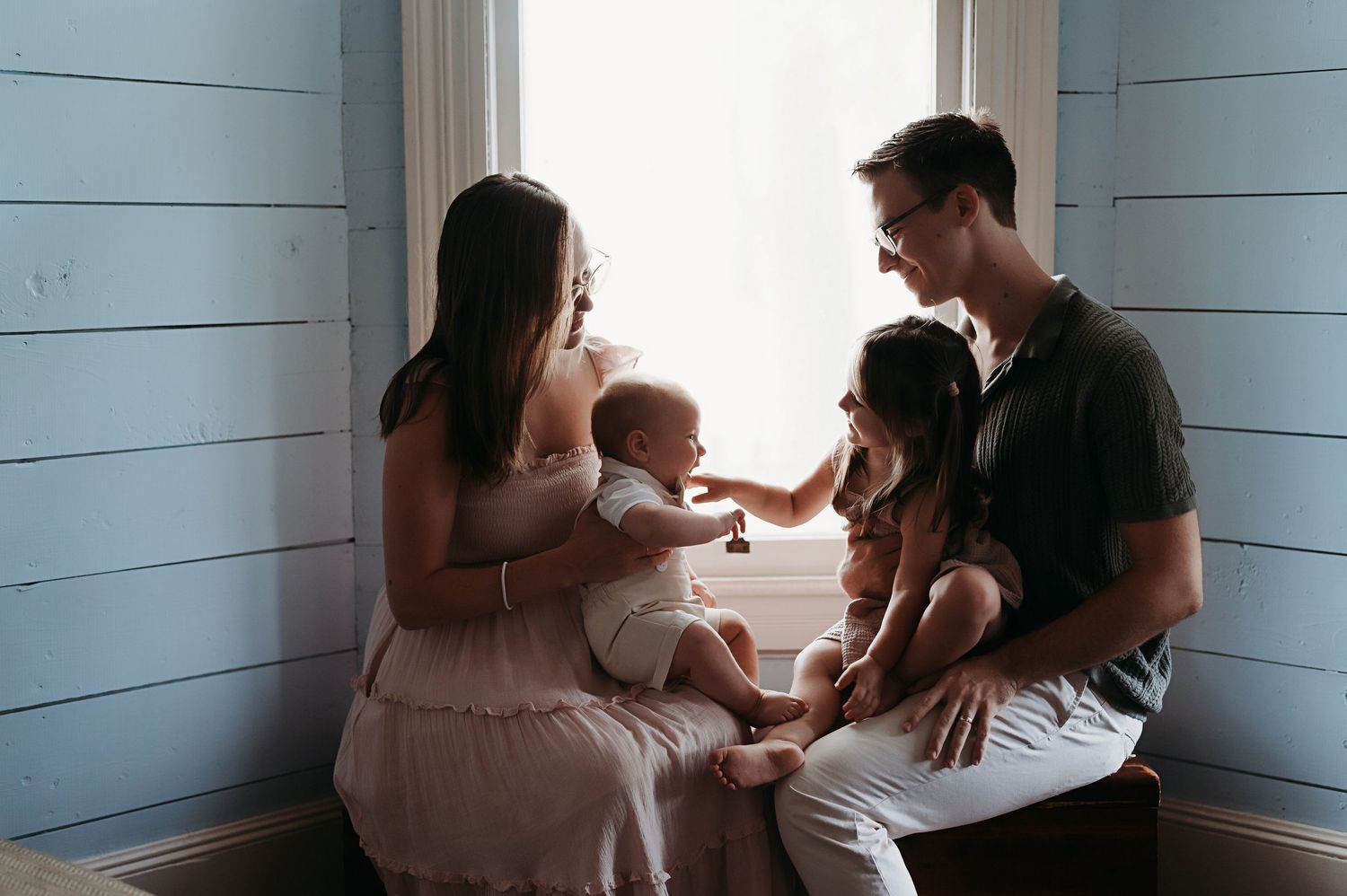 A young family with a baby and toddler shares a tender moment by a window in a cozy blue room.