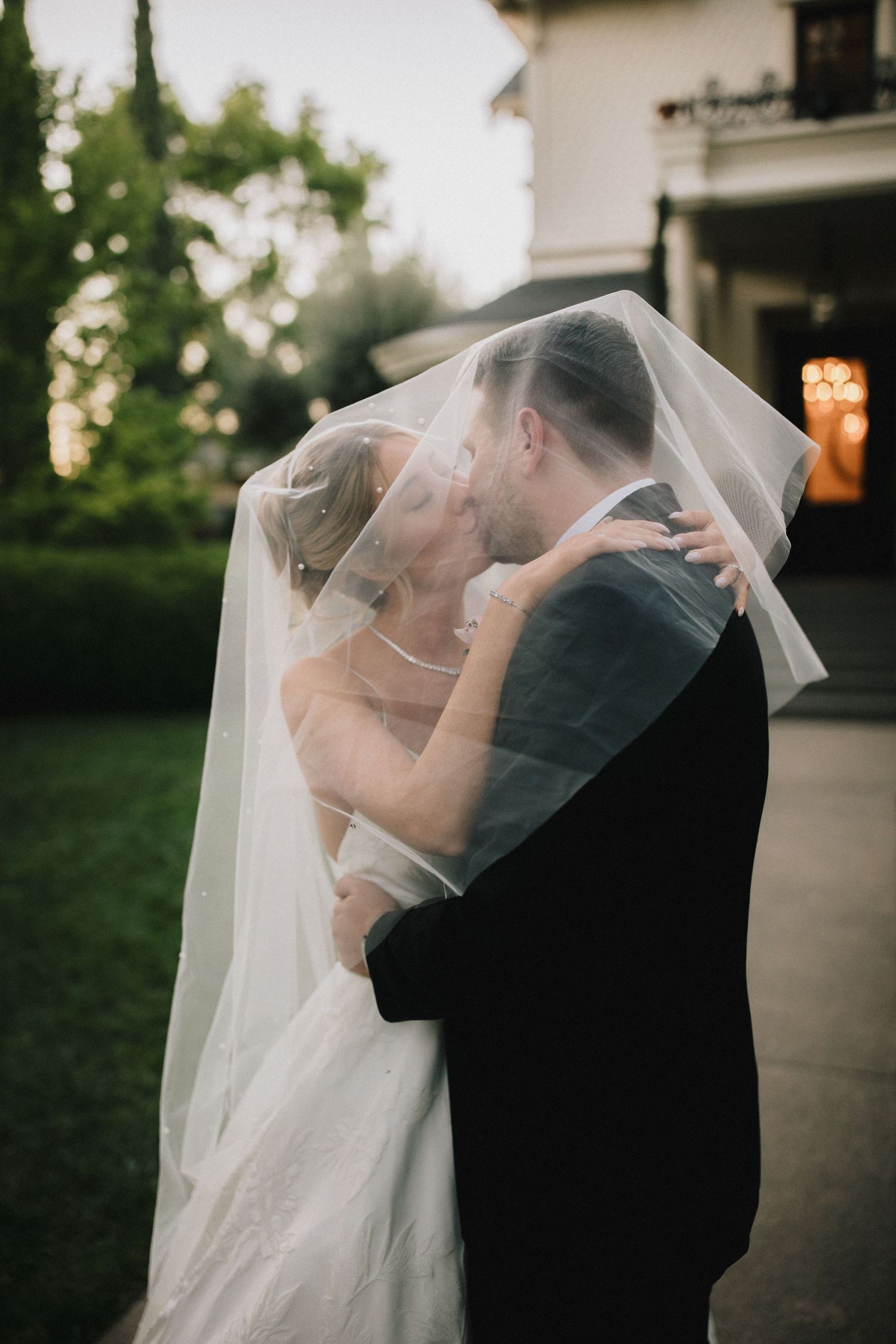 A romantic wedding moment captured outdoors with the bride in a flowing veil and groom in dark suit embracing at sunset.