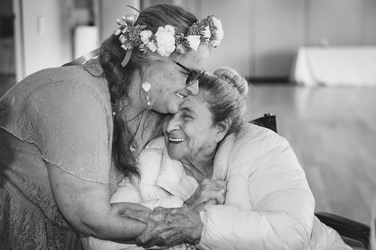 Black and white photo of two women embracing and laughing, one wearing a floral crown at a wedding celebration.