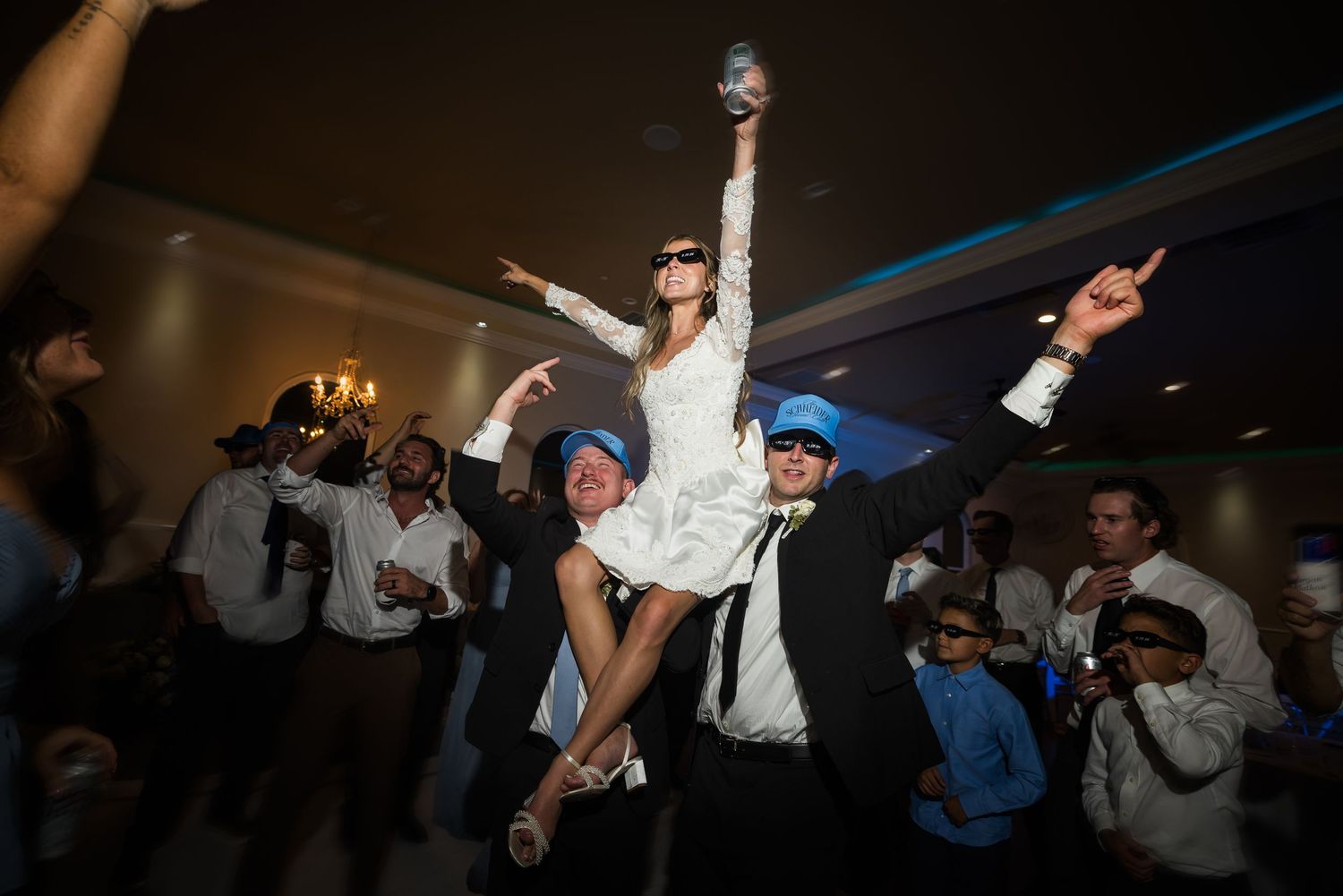 Bride lifted on shoulders during lively wedding reception dance floor celebration with guests wearing blue caps.