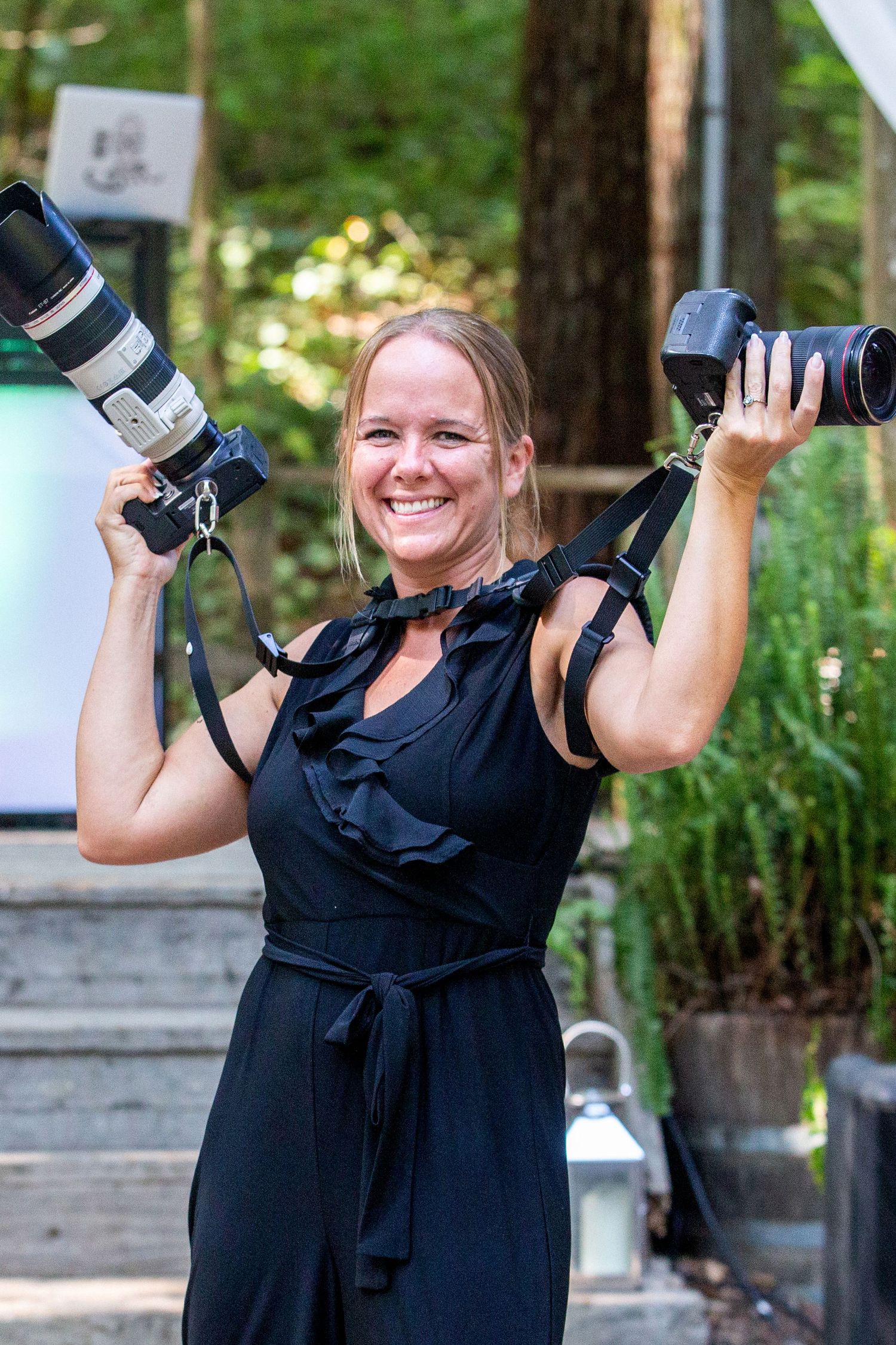 Smiling female photographer holding two cameras in a black dress outdoors in a wooded setting.