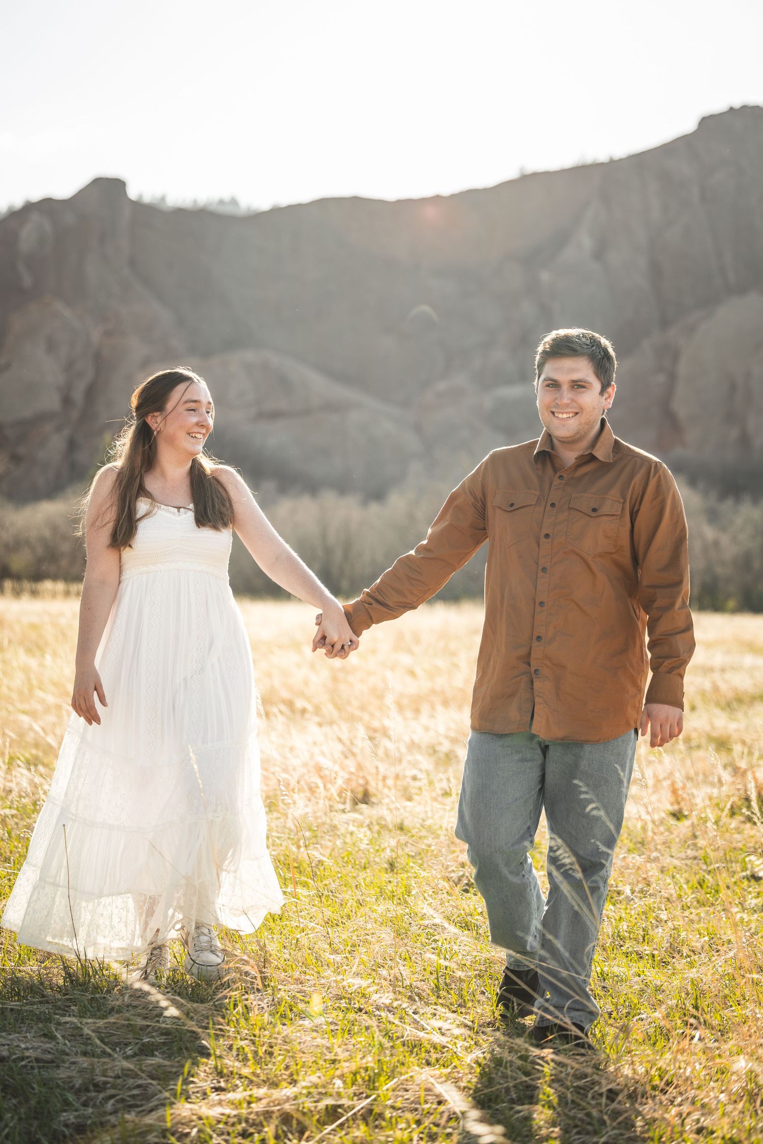 Couple holding hands walking in a sunlit field with rocky mountains in the background, woman in white dress, man in brown shirt.