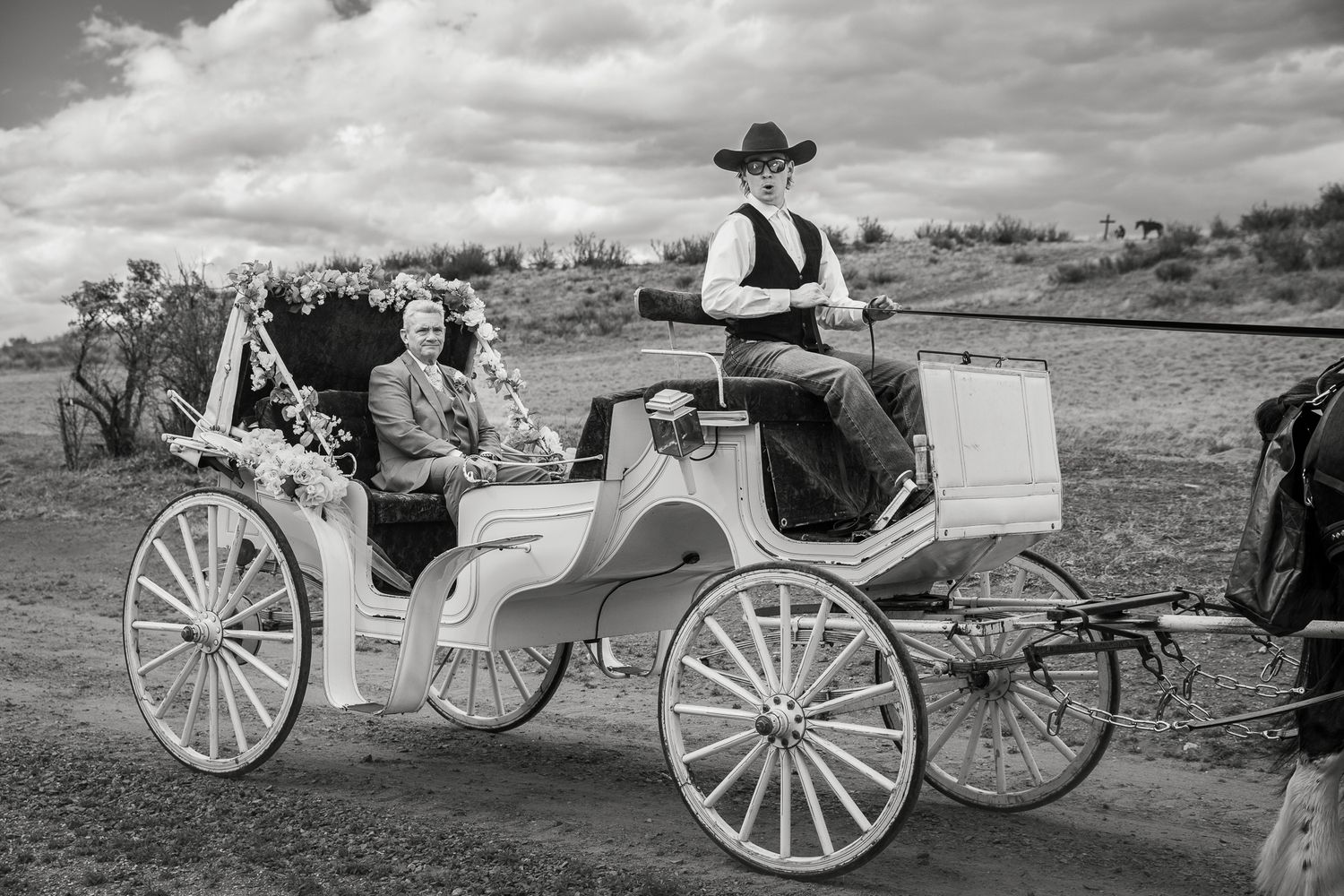 Black and white photo of a couple riding in an elegant white horse-drawn carriage driven by a man in a cowboy hat.