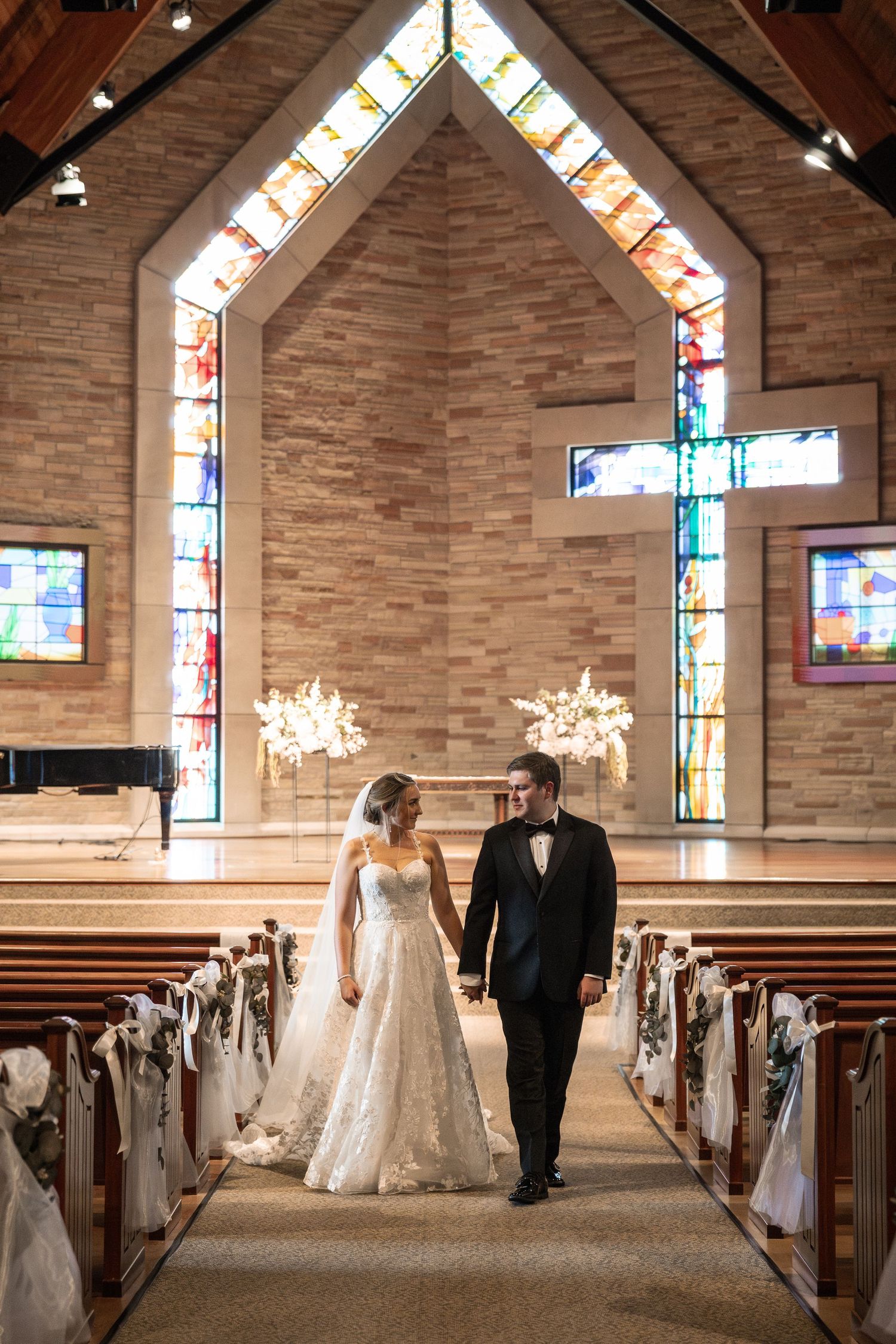 Bride and groom walking hand in hand down church aisle with stained glass windows and wooden cross backdrop.