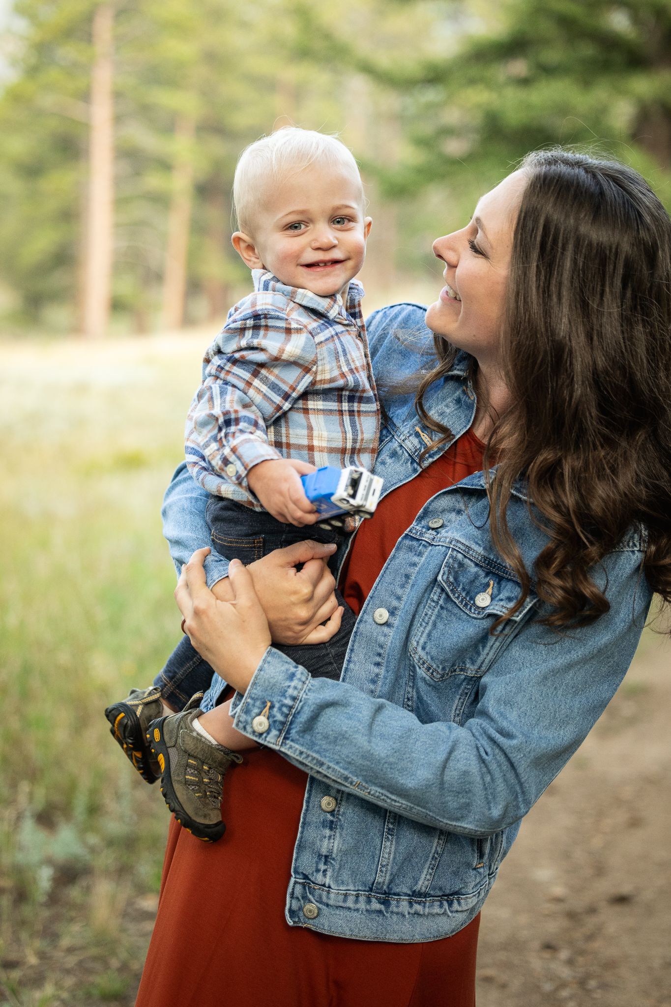A smiling mother in a denim jacket holds her happy baby boy outdoors on a nature path surrounded by trees.