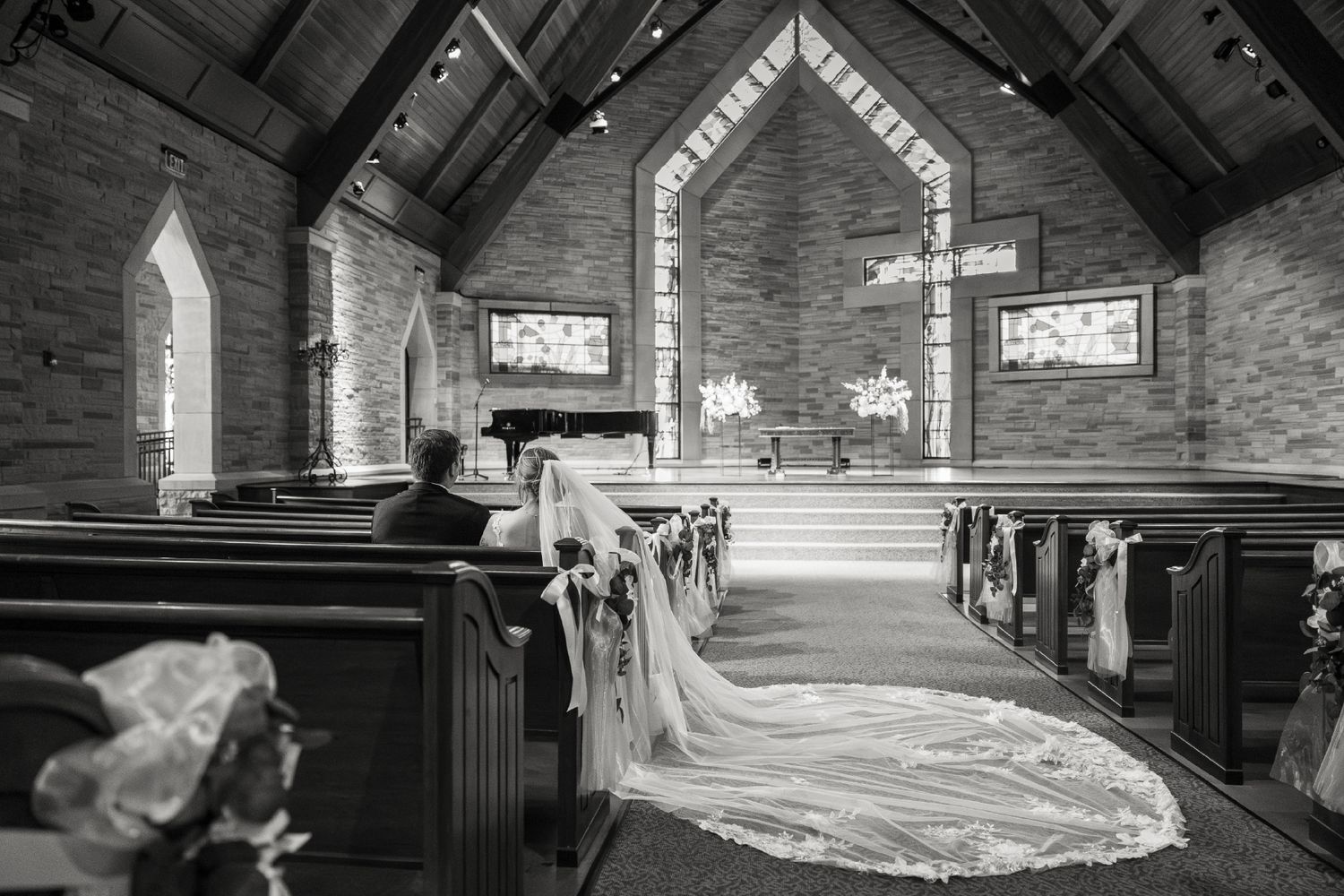 Black and white wedding photo of a church interior with pews, floral decorations, and a long bridal veil train along the aisle.