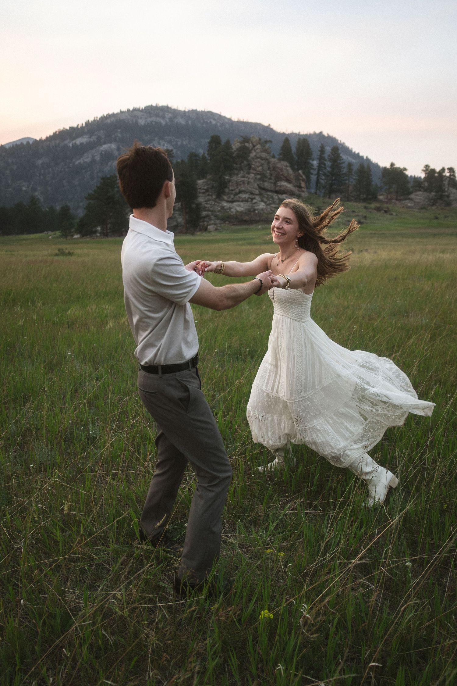 Couple dancing joyfully in a green meadow at dusk, woman in white dress spinning, mountains in background.