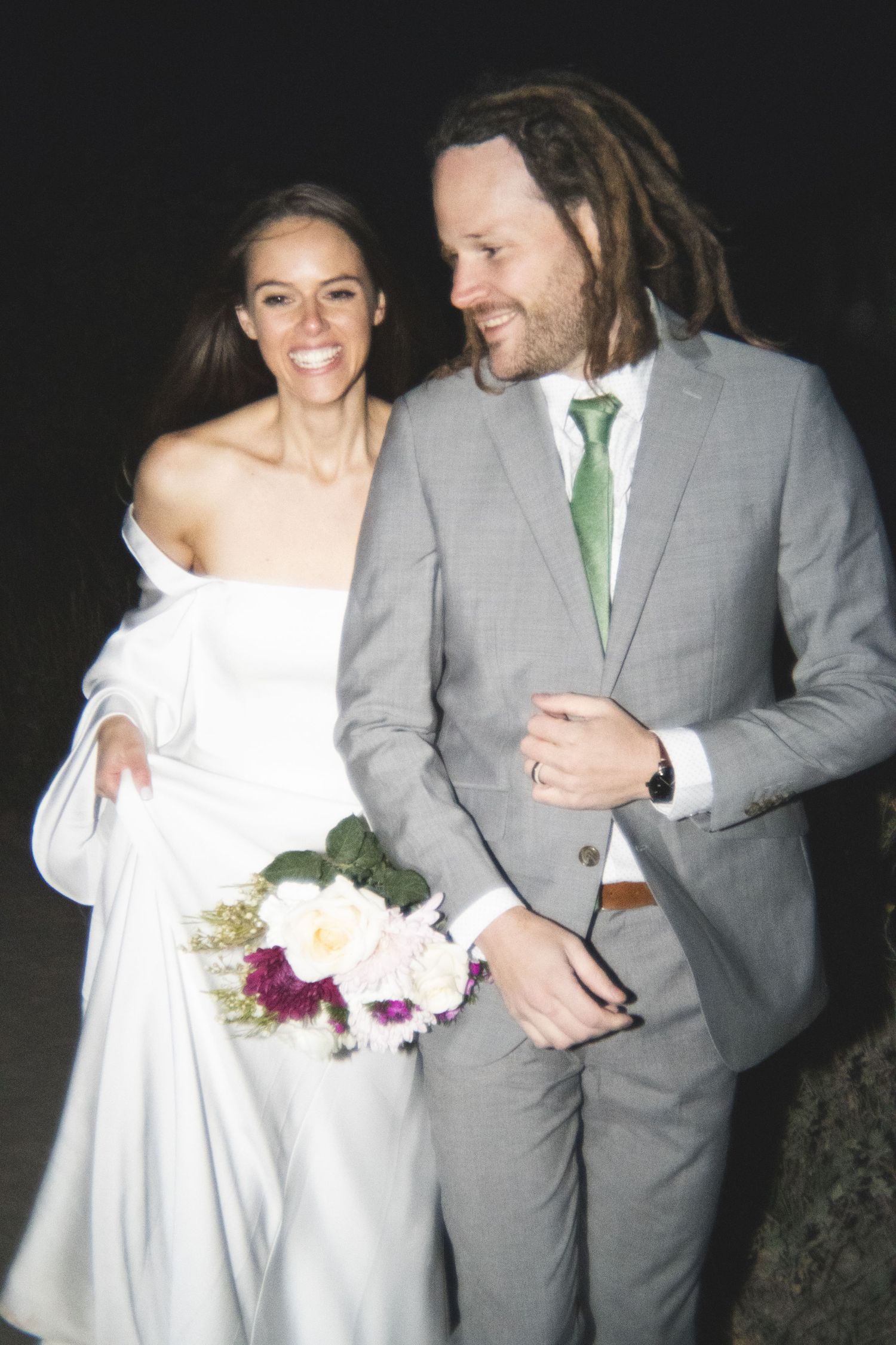 A joyful wedding couple laughs together, with the groom in a grey suit and green tie.