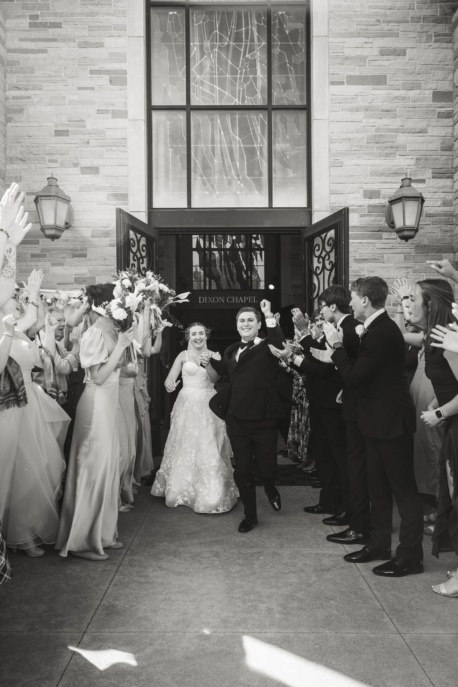 Bride and groom exit church doors through cheering guests in black and white wedding ceremony sendoff photo.