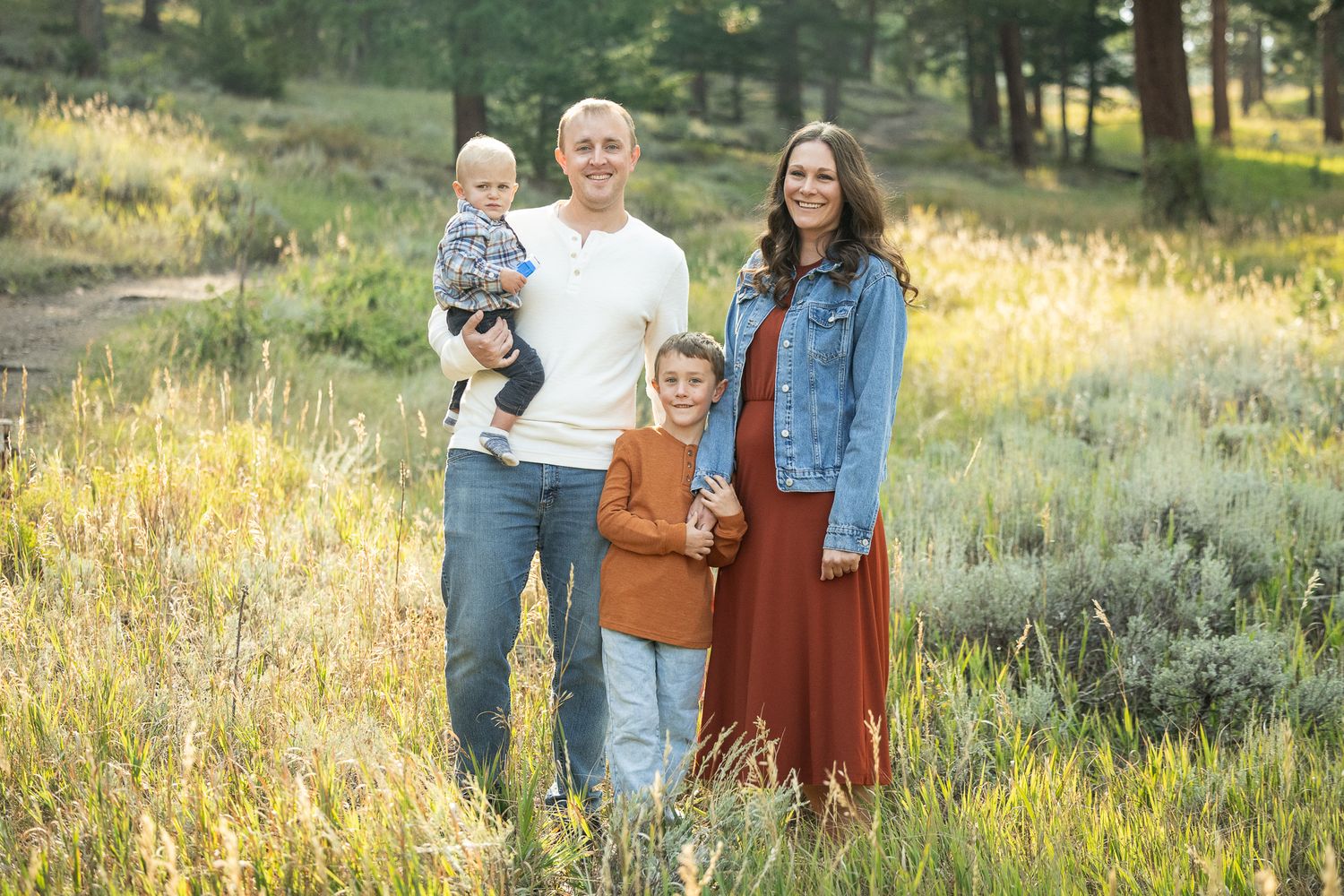 Family of four posing outdoors in a meadow, parents with two young children, wearing casual fall outfits in warm earth tones.