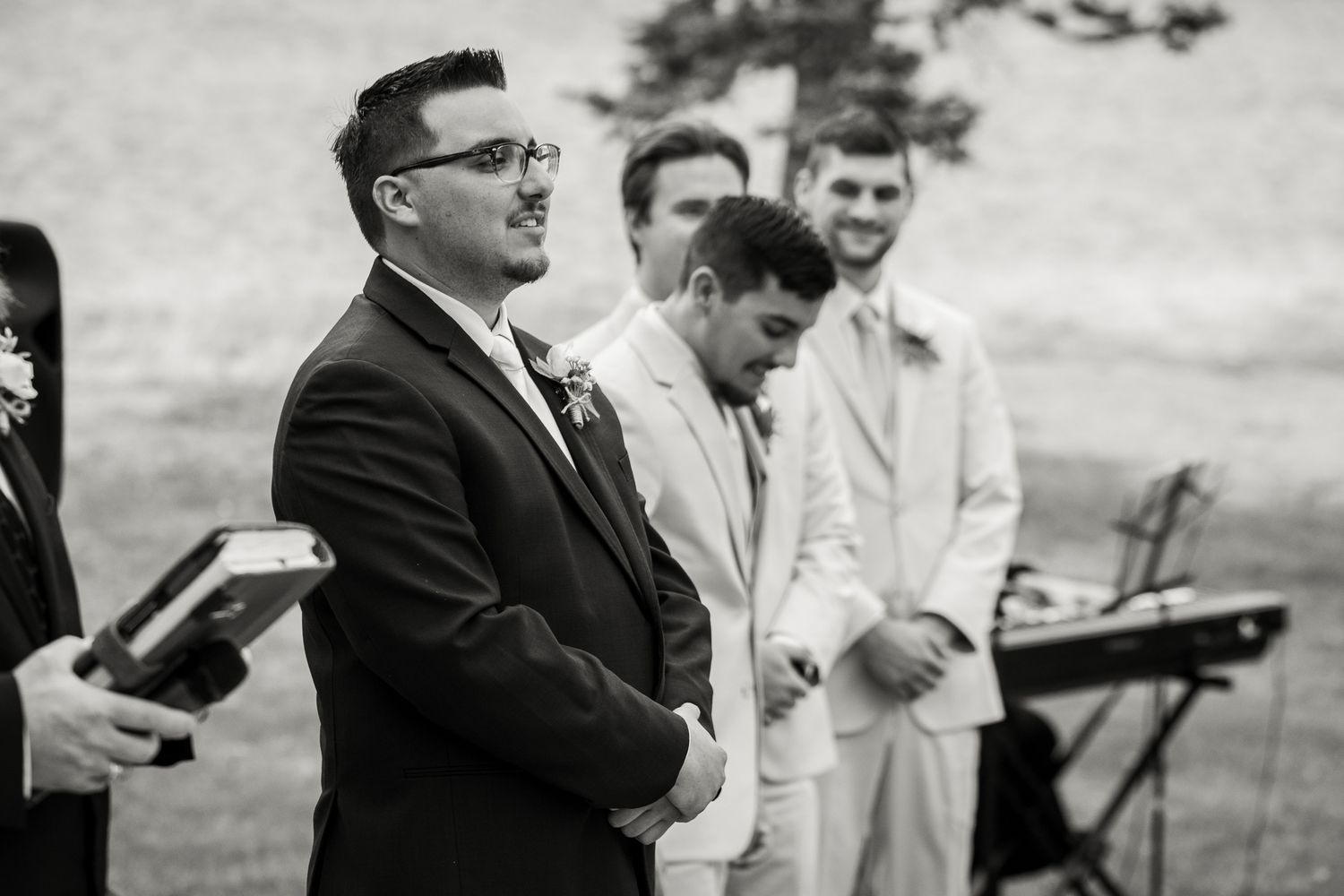 Black and white photo of groom watching bride walk down aisle at outdoor wedding ceremony with groomsmen standing behind.
