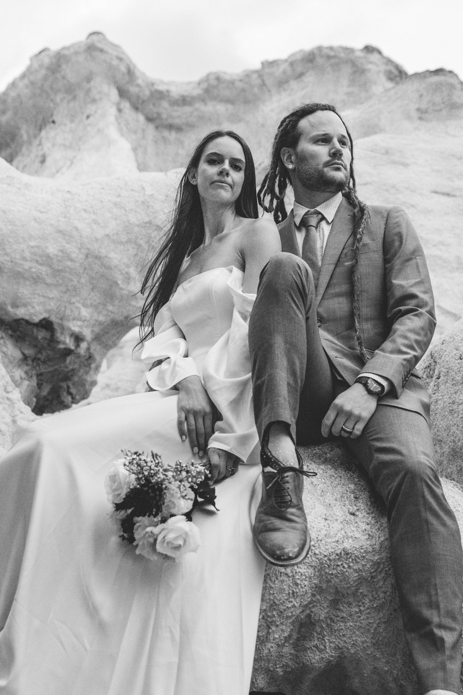 A black and white portrait of newlyweds sitting on rocky terrain.