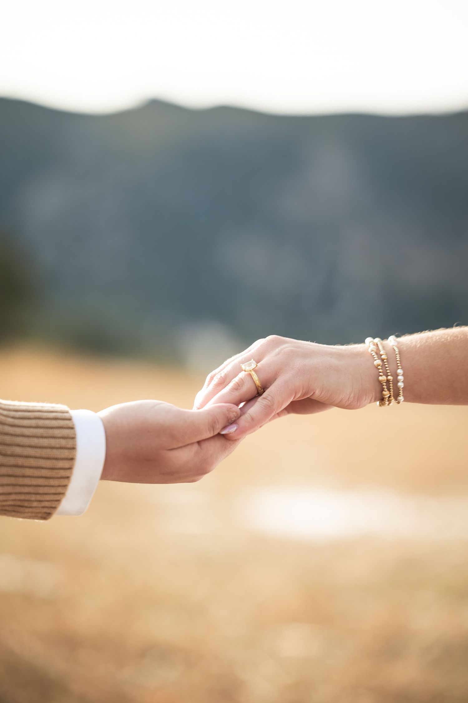 Two hands gently holding each other outdoors, one adorned with gold bracelets and a ring, with a blurred mountain backdrop.