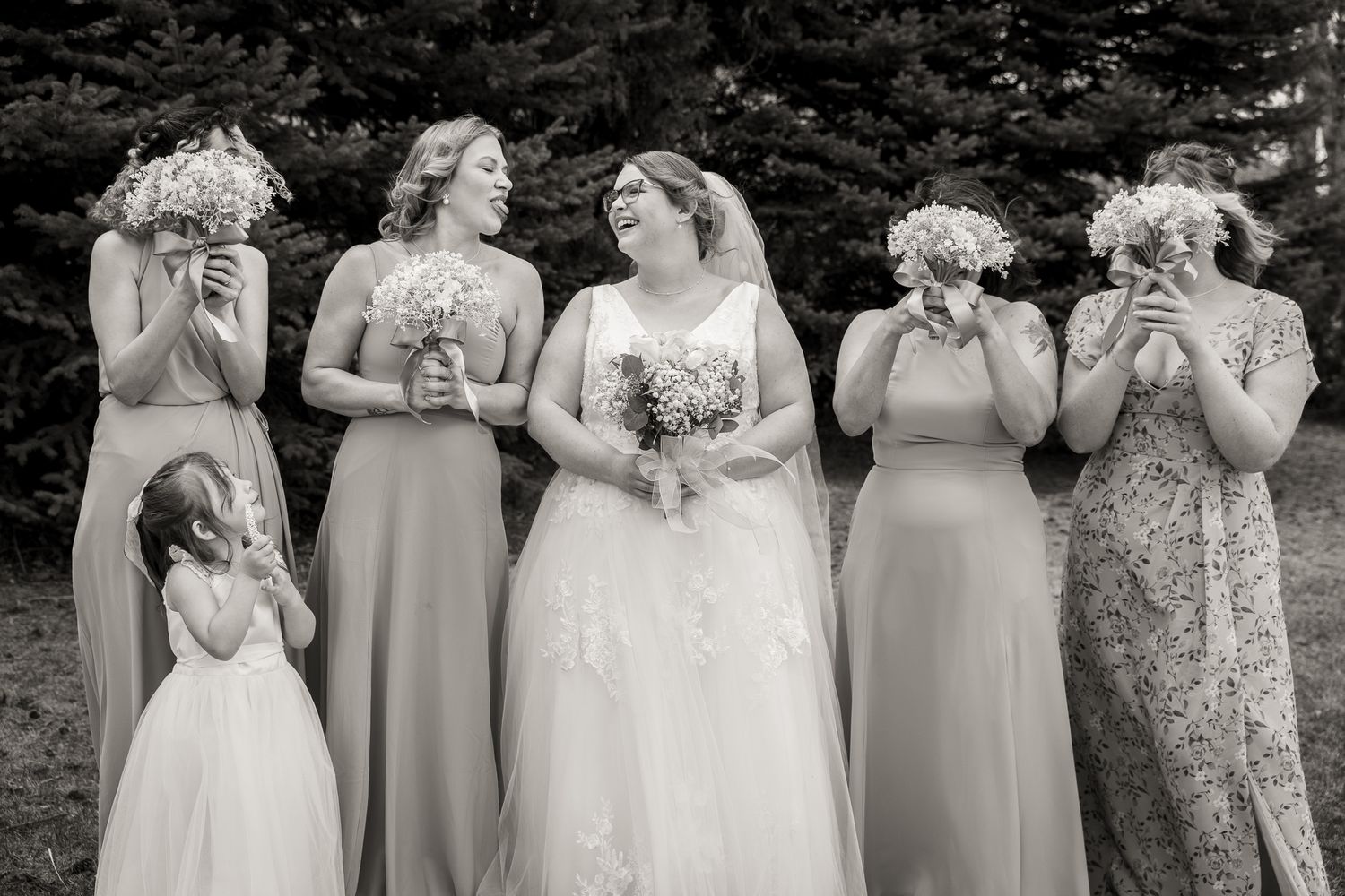 Black and white photo of bride with bridesmaids holding bouquets, some covering faces, and a flower girl at a wedding.
