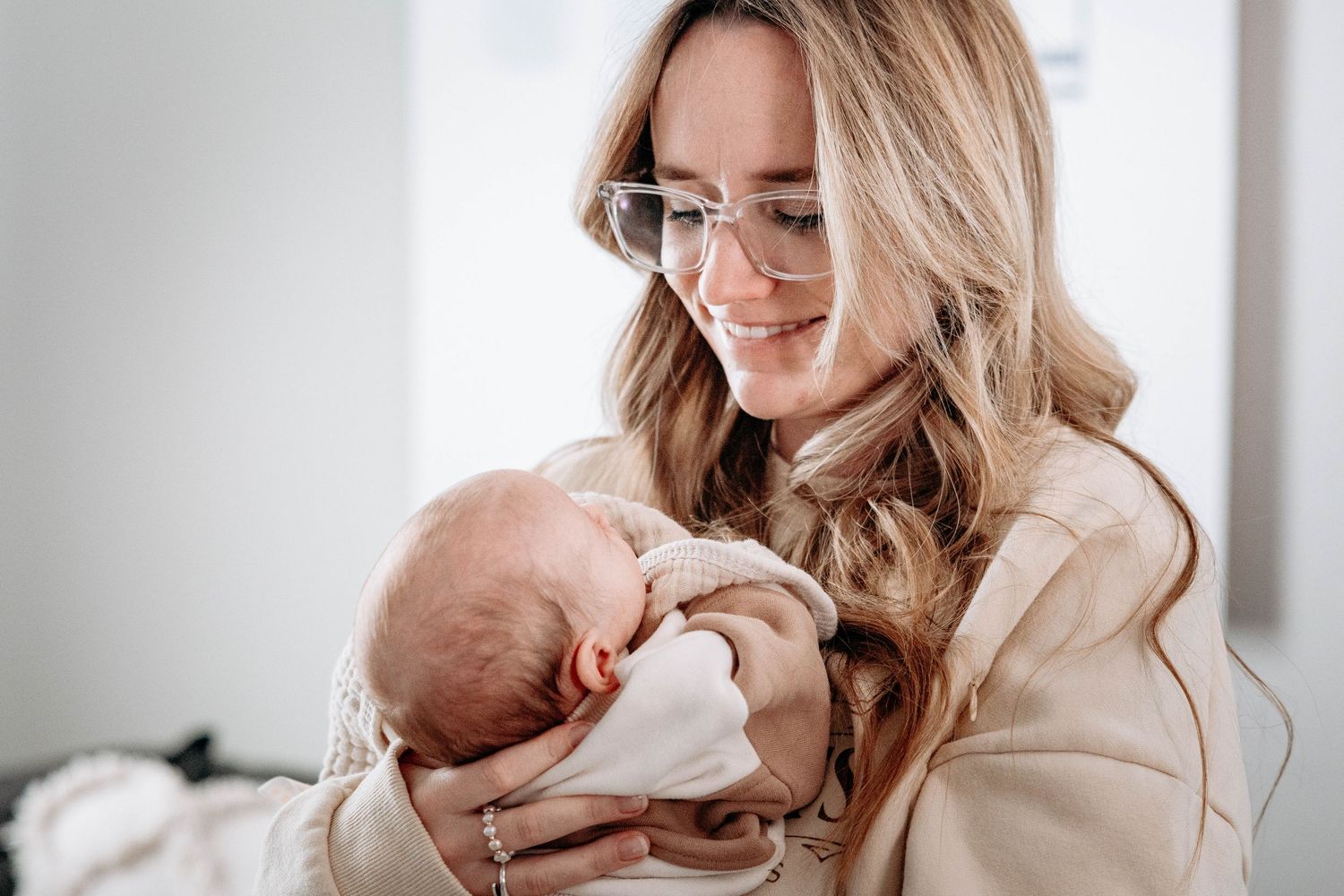 A smiling woman with glasses gently cradles a sleeping newborn baby in her arms.