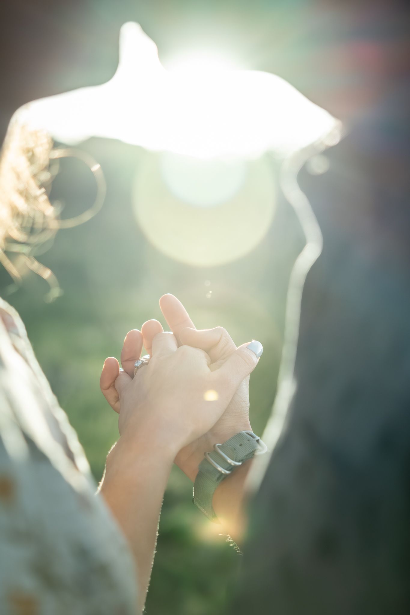 Couple holding hands with engagement ring visible, bathed in warm sunlight flare outdoors.