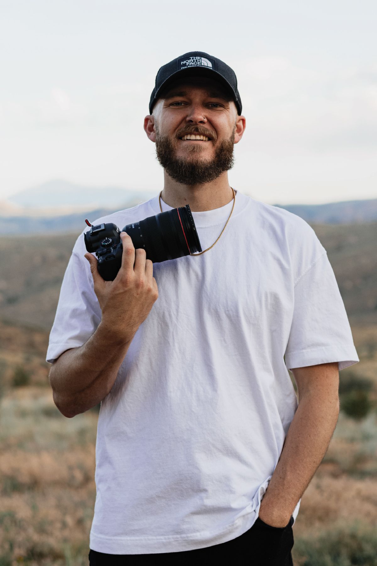 Smiling bearded man holding a DSLR camera outdoors, wearing a white t-shirt and black North Face cap.
