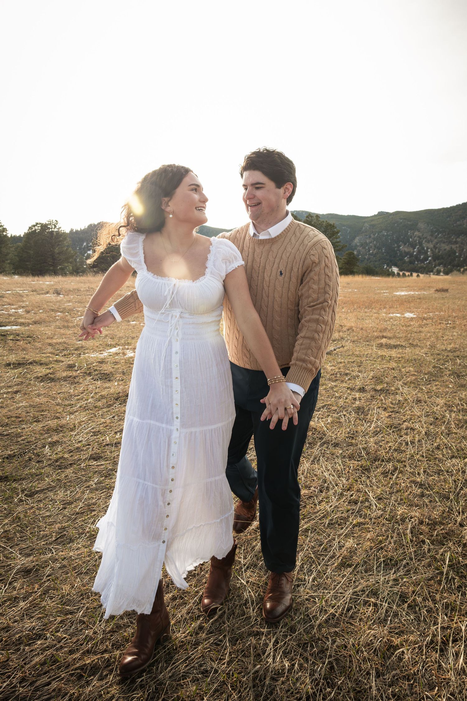 Couple walking hand-in-hand in a sunlit field, woman in white dress and boots, man in tan sweater, mountains behind.