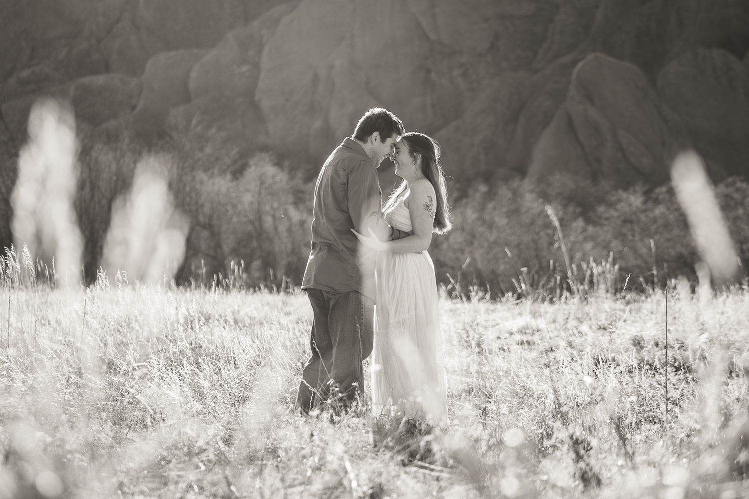 Black and white photo of a romantic couple embracing in a sunlit field with rocky mountains in the background.