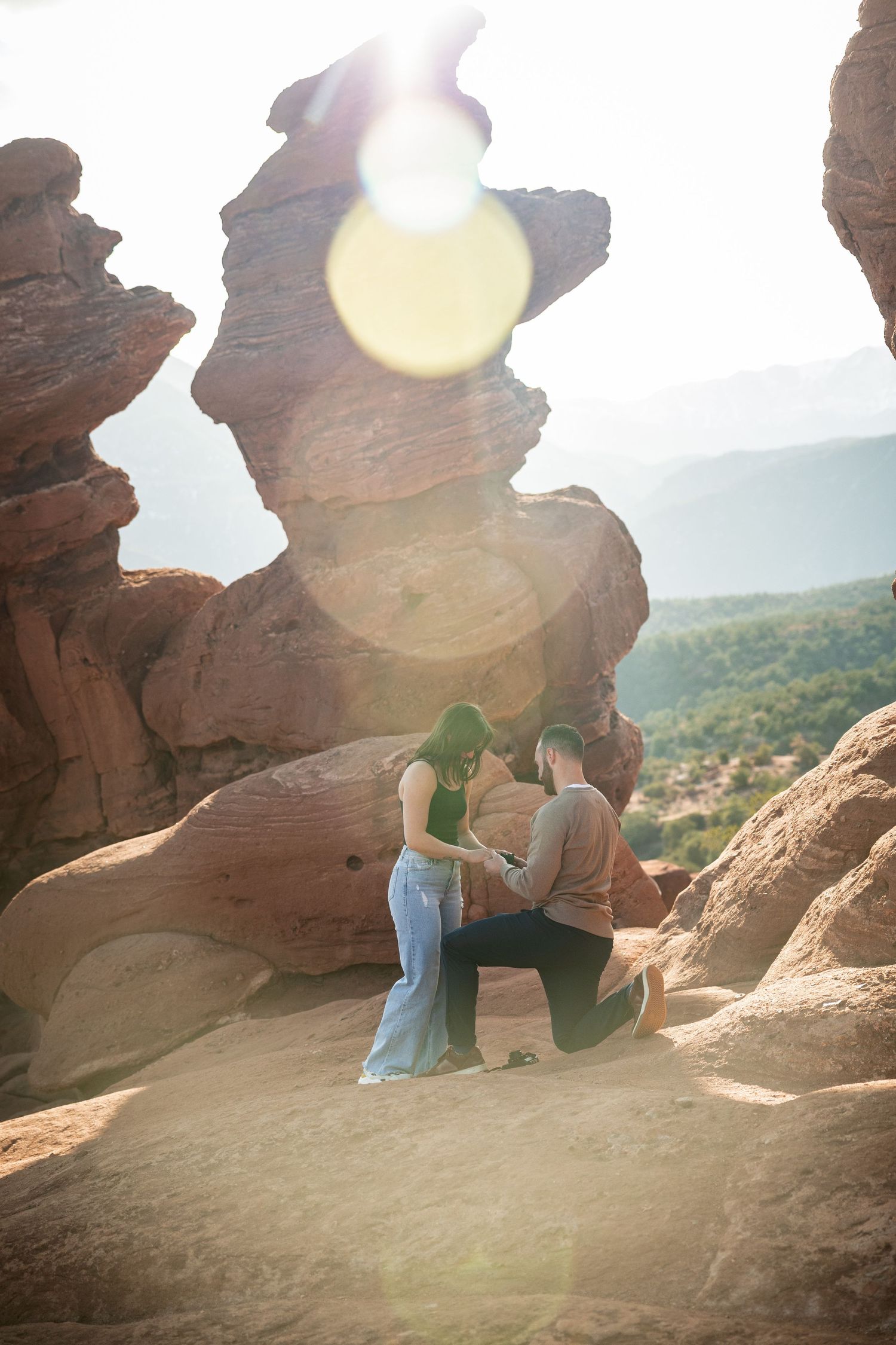 Couple sitting on red rock formations with sun flare and valley views.