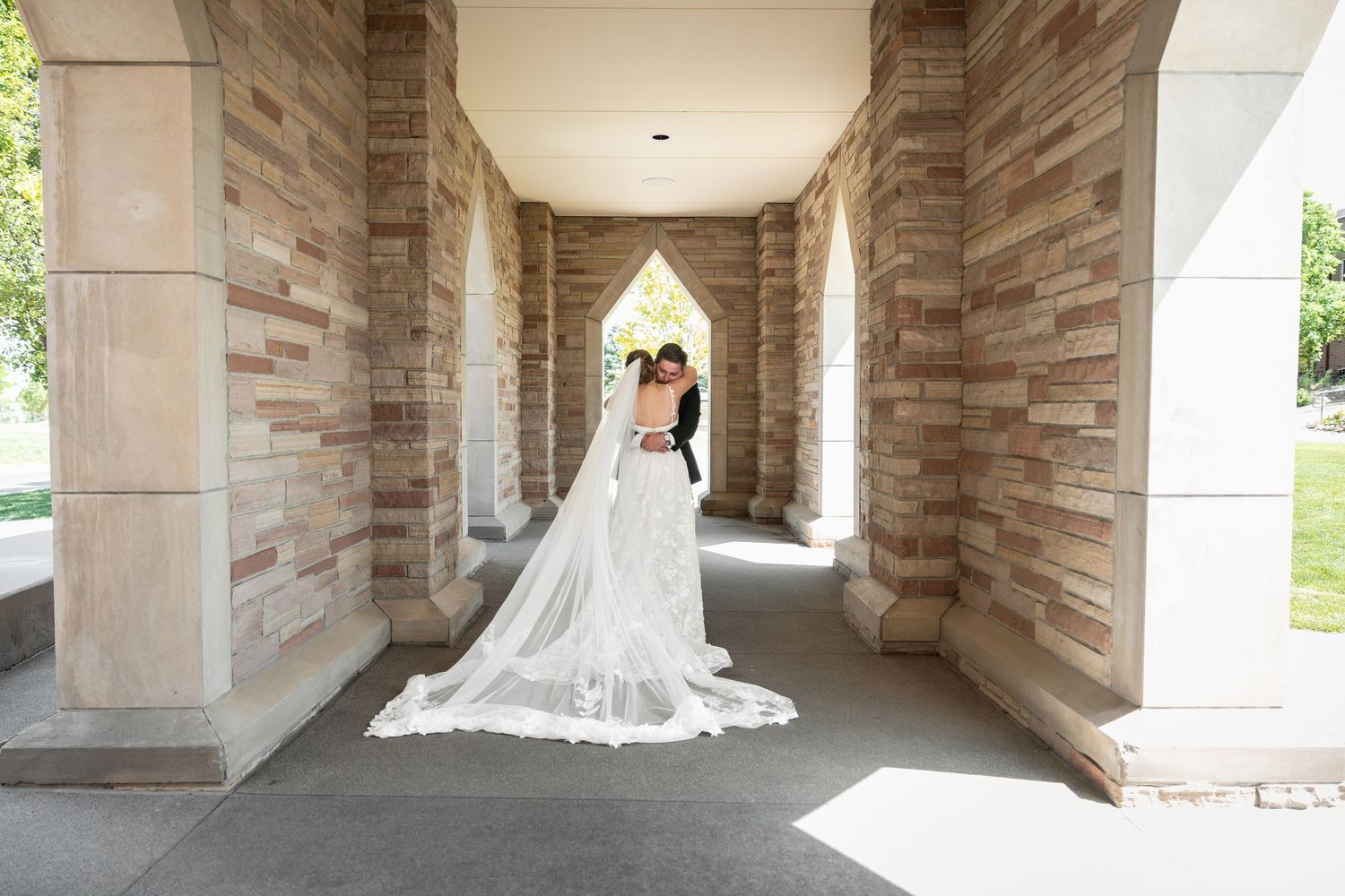 Bride and groom embrace in a stunning stone archway corridor, with a long flowing wedding veil trailing on the ground.