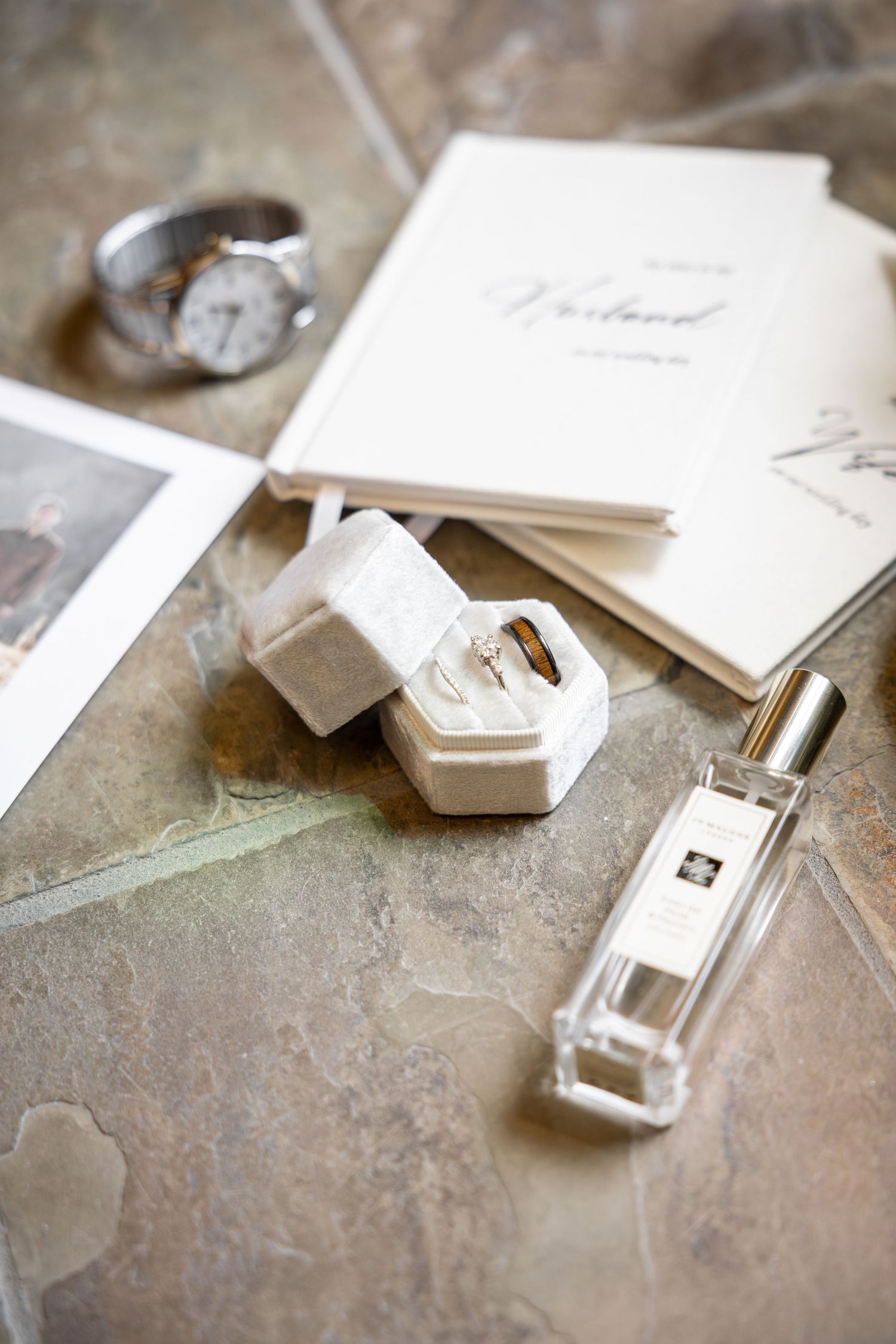 Elegant wedding flat lay with engagement ring in velvet box, Jo Malone perfume, watch, and stationery on stone surface.