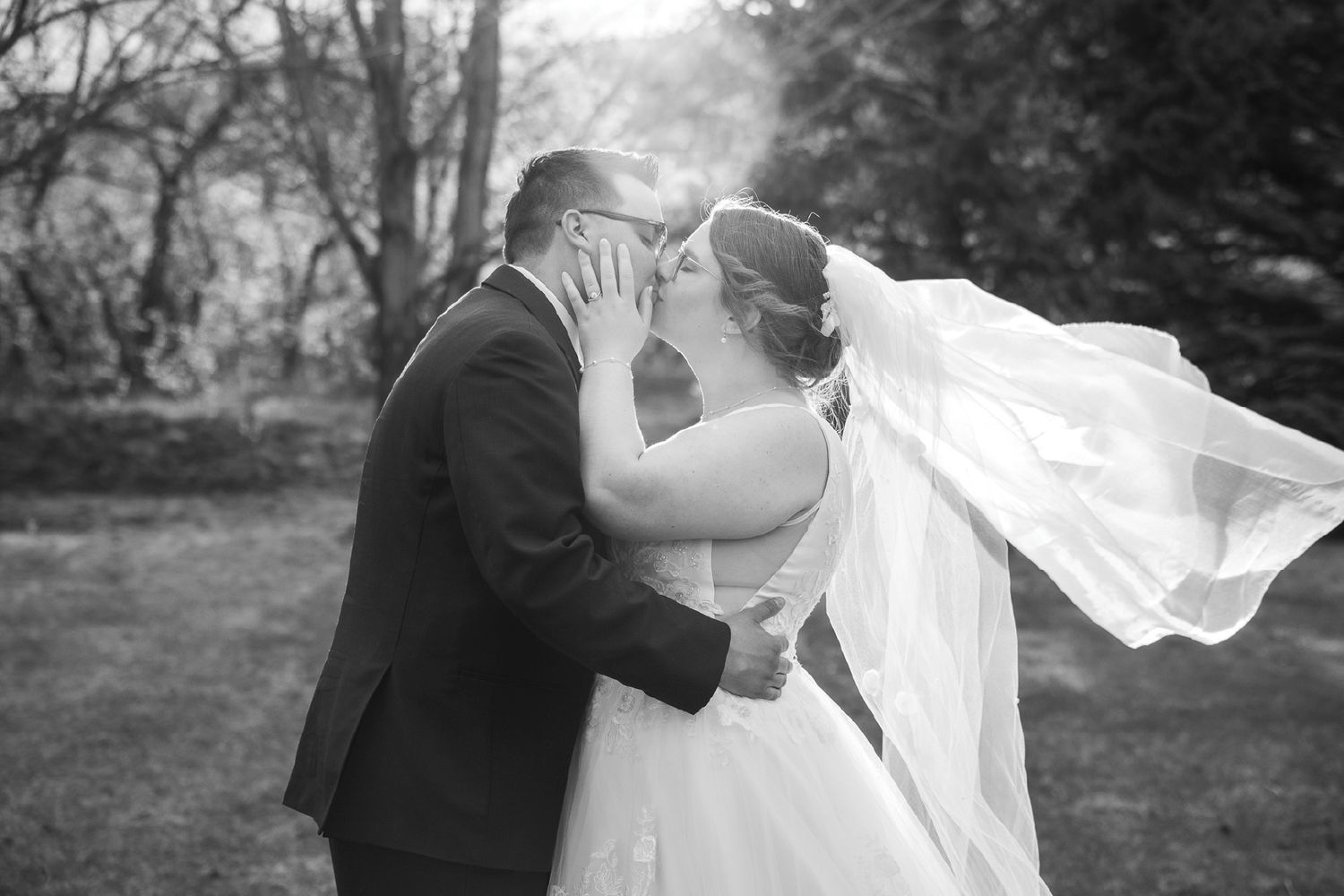Black and white photo of a bride and groom sharing a kiss outdoors, with the bride's veil flowing in the wind.