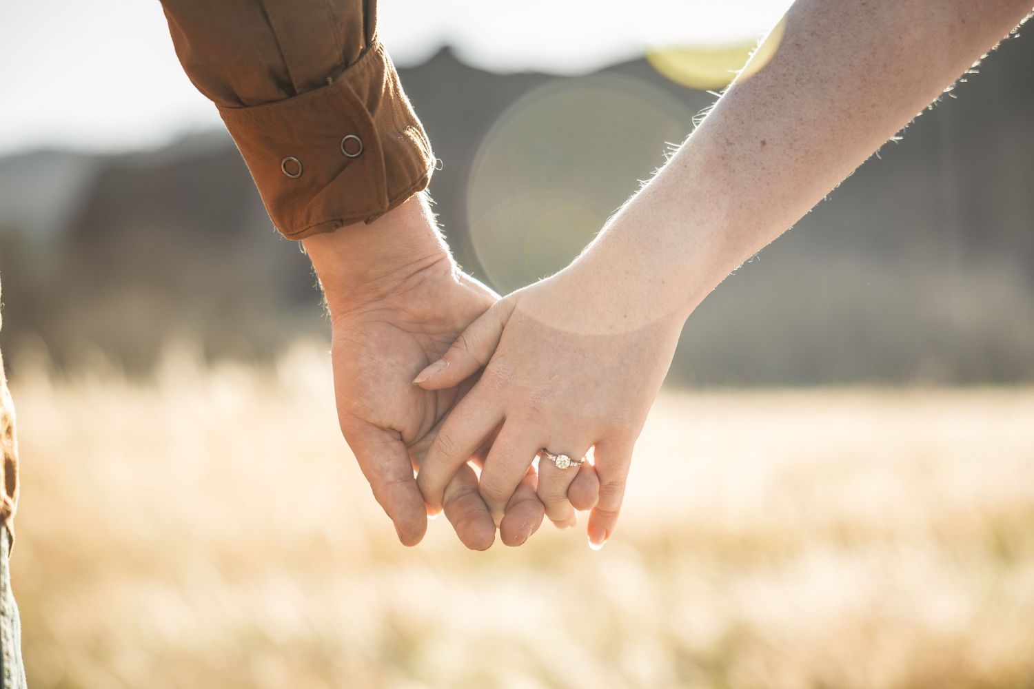 Couple holding hands in a golden field, woman wearing a diamond engagement ring, romantic outdoor portrait.