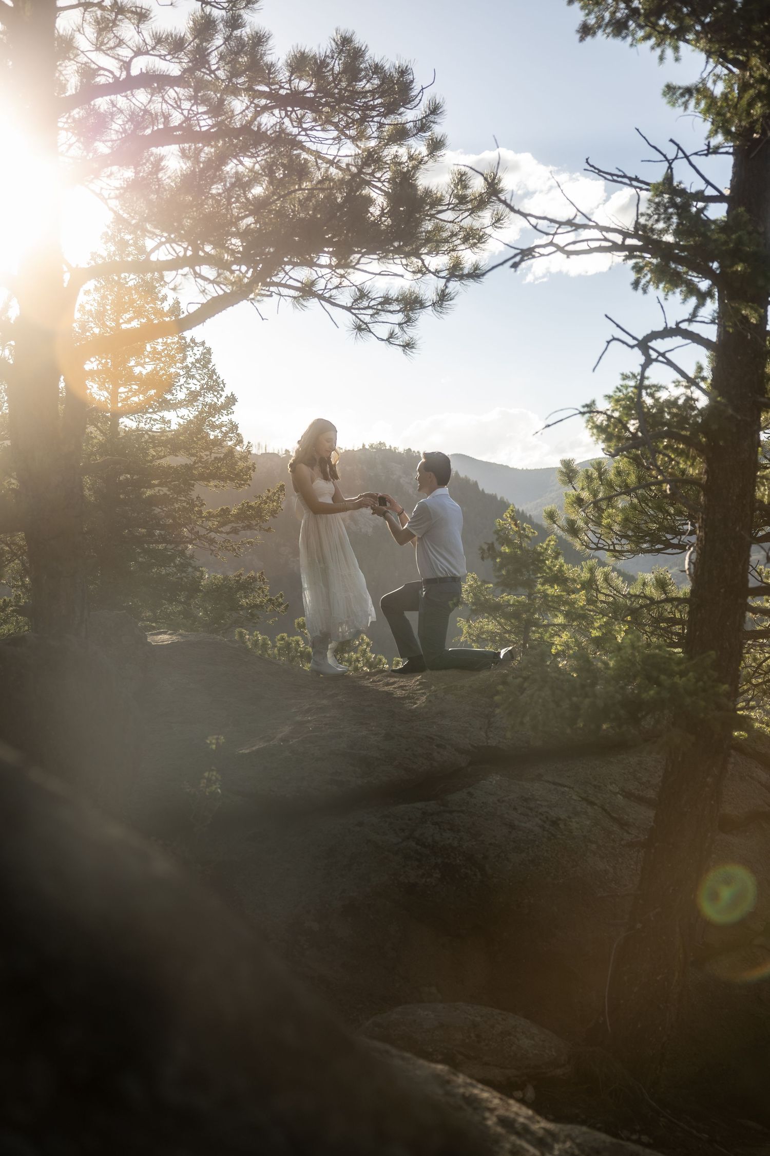 Man kneeling on rocky overlook proposing to woman in white dress among pine trees with golden sunlight and mountain views.
