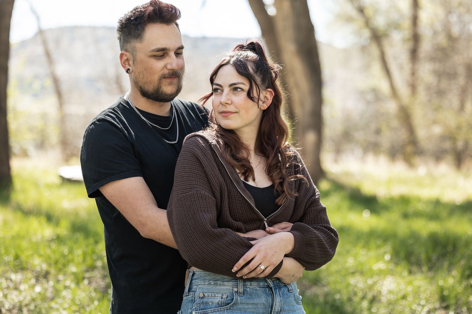 A couple embraces outdoors in a sunny park, the man holding the woman from behind among green trees.