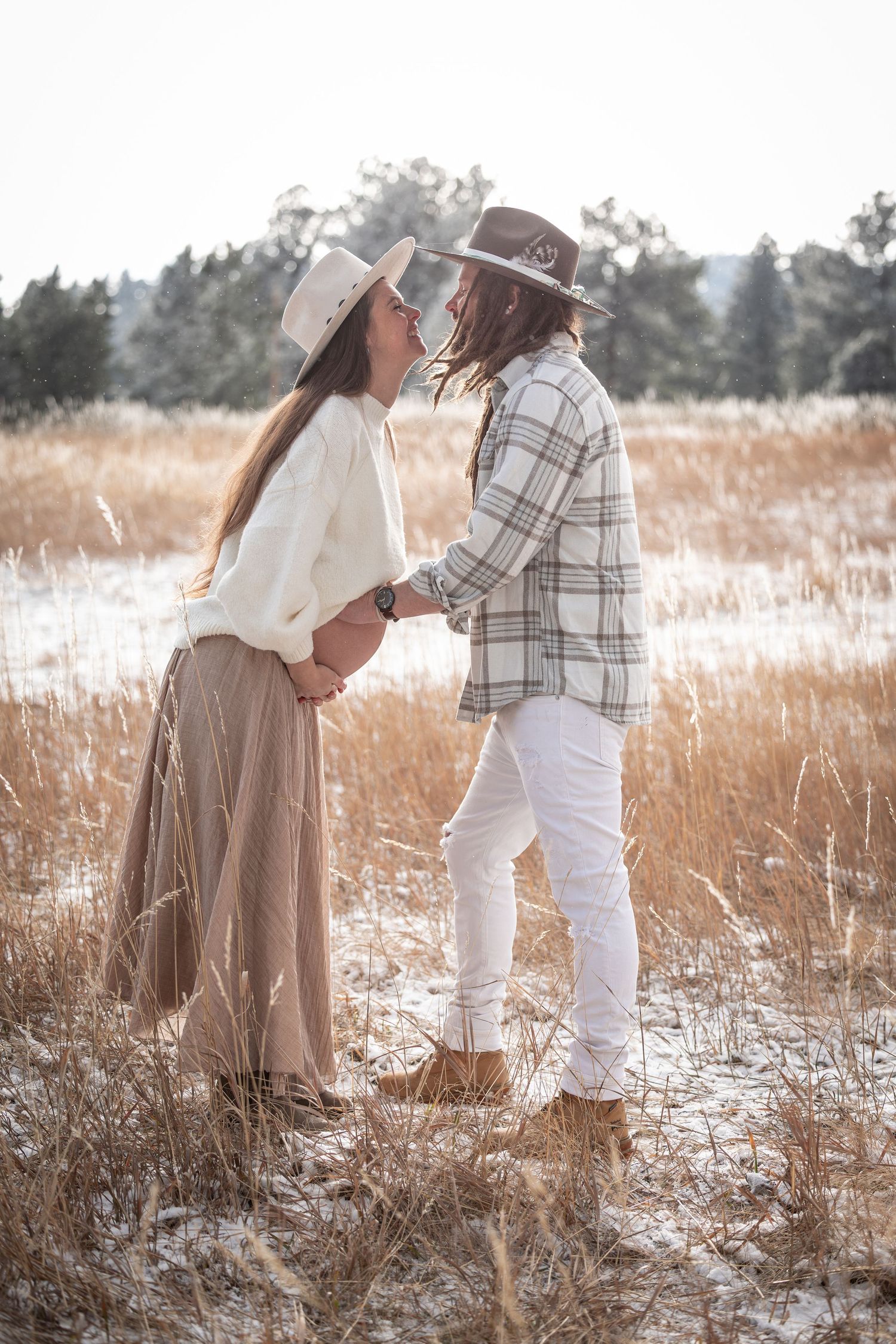 A couple in white and beige outfits embraces in a snowy winter field with evergreen trees in the background.