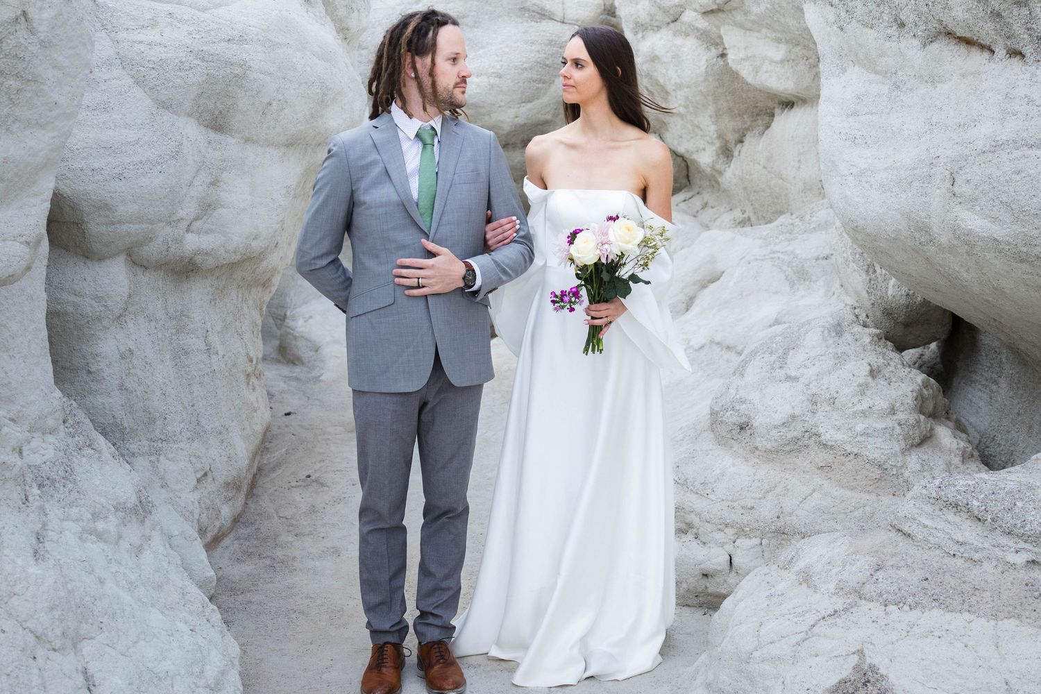 Wedding couple stands against white rock formations in formal attire with bouquet.