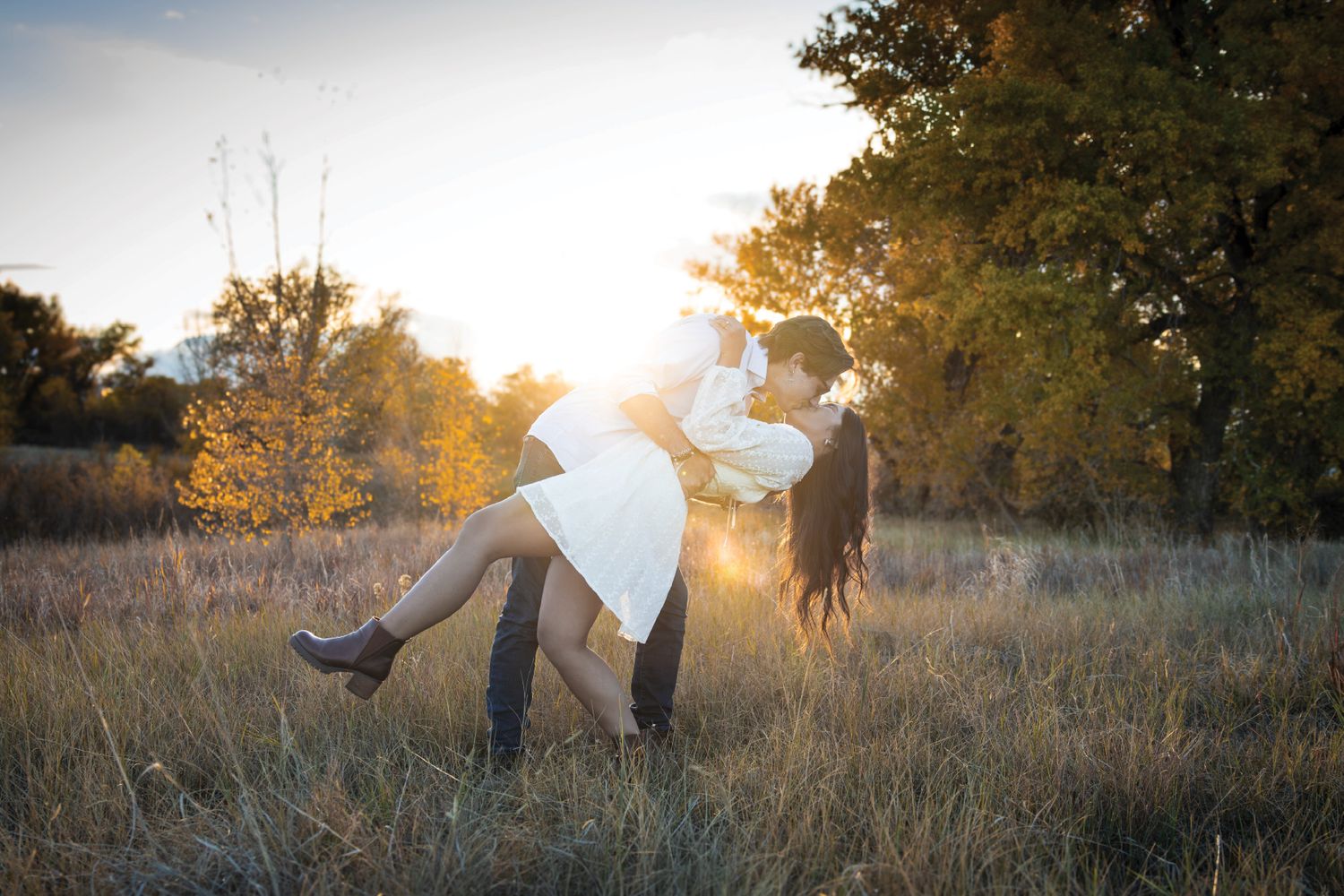 Couple sharing a romantic kiss in a sunlit field during golden hour with autumn trees in the background.