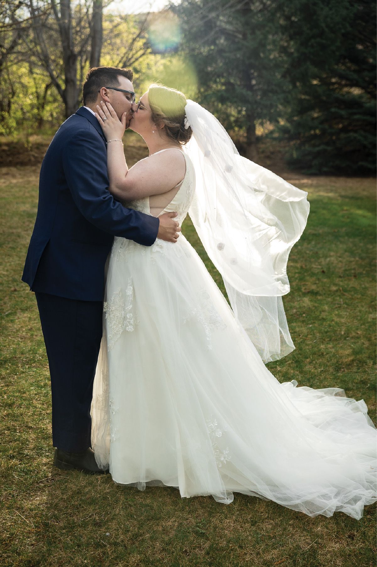 Bride and groom share a romantic kiss outdoors, her white gown and veil flowing in the breeze on their wedding day.