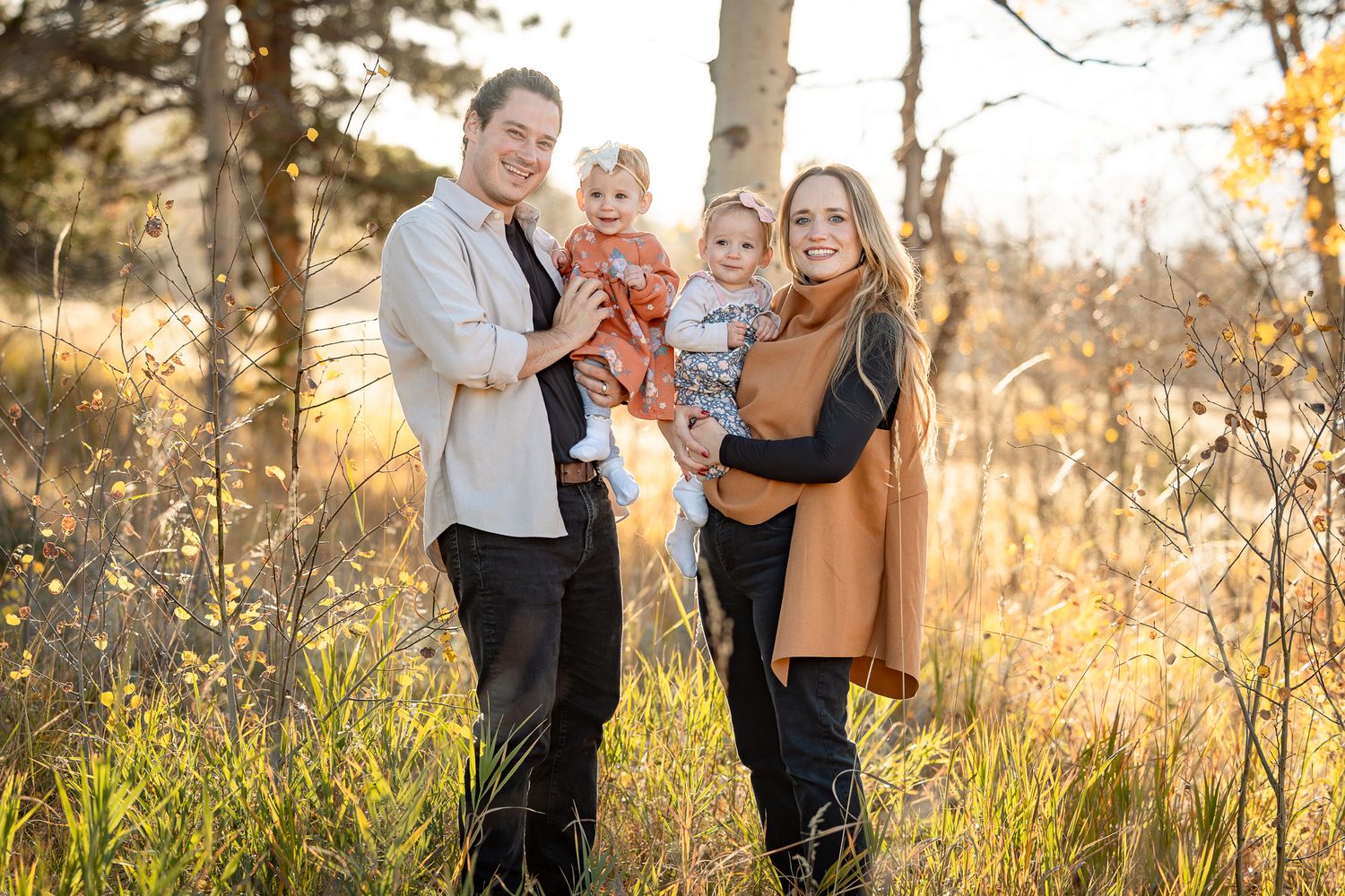 Family of four poses outdoors in golden autumn light, parents holding two young children among tall grasses and trees.