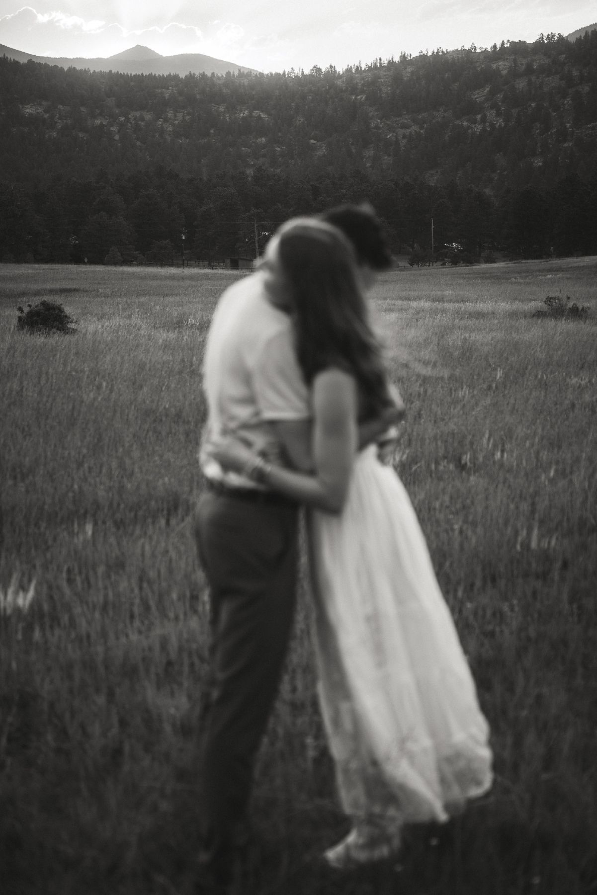 Black and white photo of a couple embracing in a meadow with mountains and forest in the background.